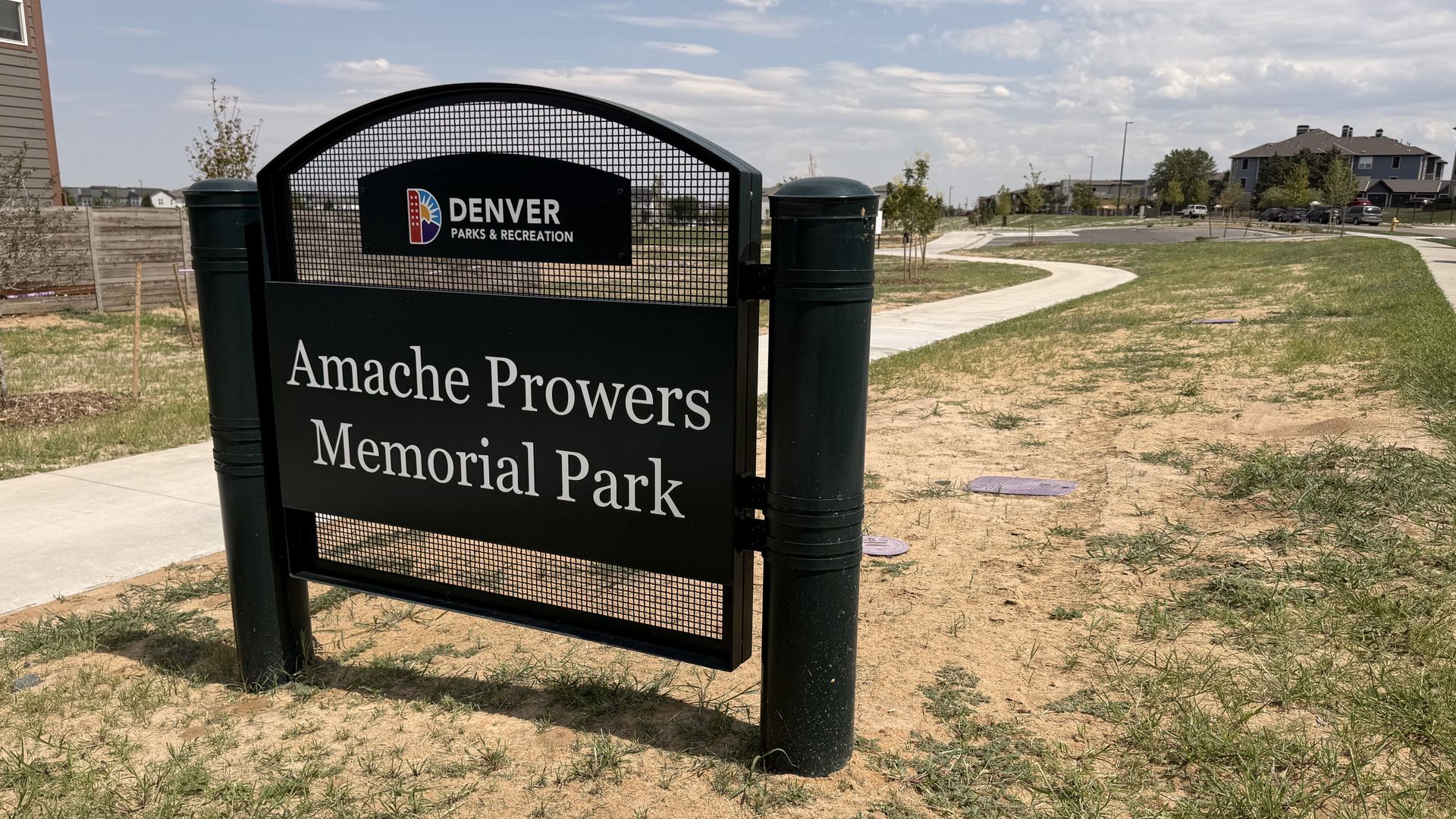 Foreground shows a dark green Denver Parks & Recreation sign with mesh, stating "Amache Prowers Memorial Park"; a curved path, dry grass, and a suburban street with houses under a blue sky.