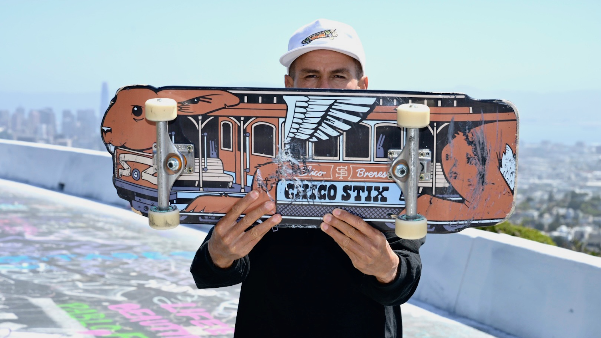 Photo of Chico Brenes holding a skateboard marked with graphics toward the camera against the Twin Peaks backdrop
