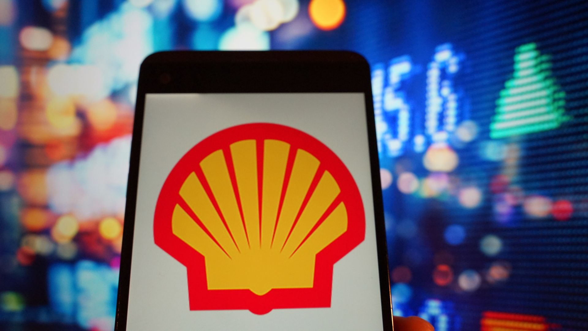 In this image, a phone with the Shell logo is held up to the camera. Lights and numbers are in the background.