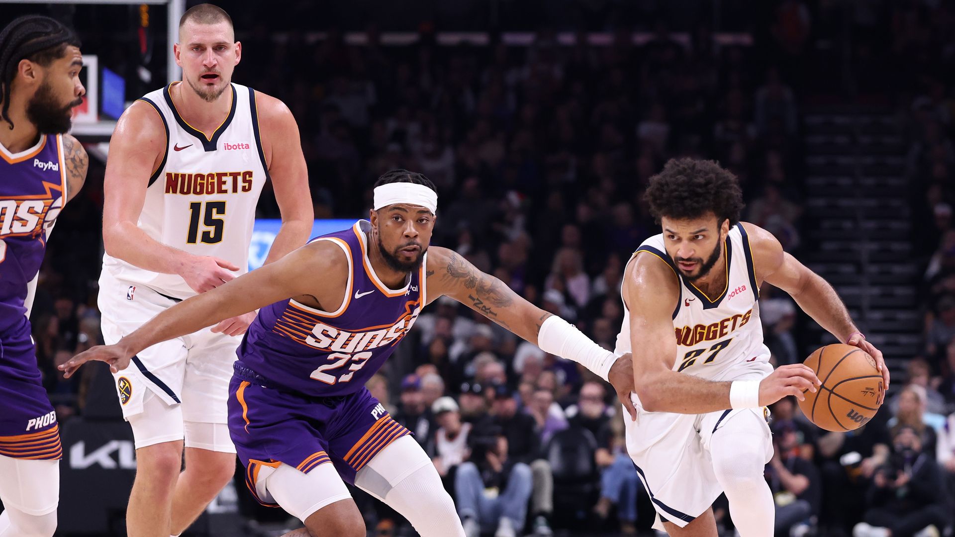 Denver Nuggets player dribbles basketball while Phoenix Suns defender tries to block, other players in background on basketball court during game.