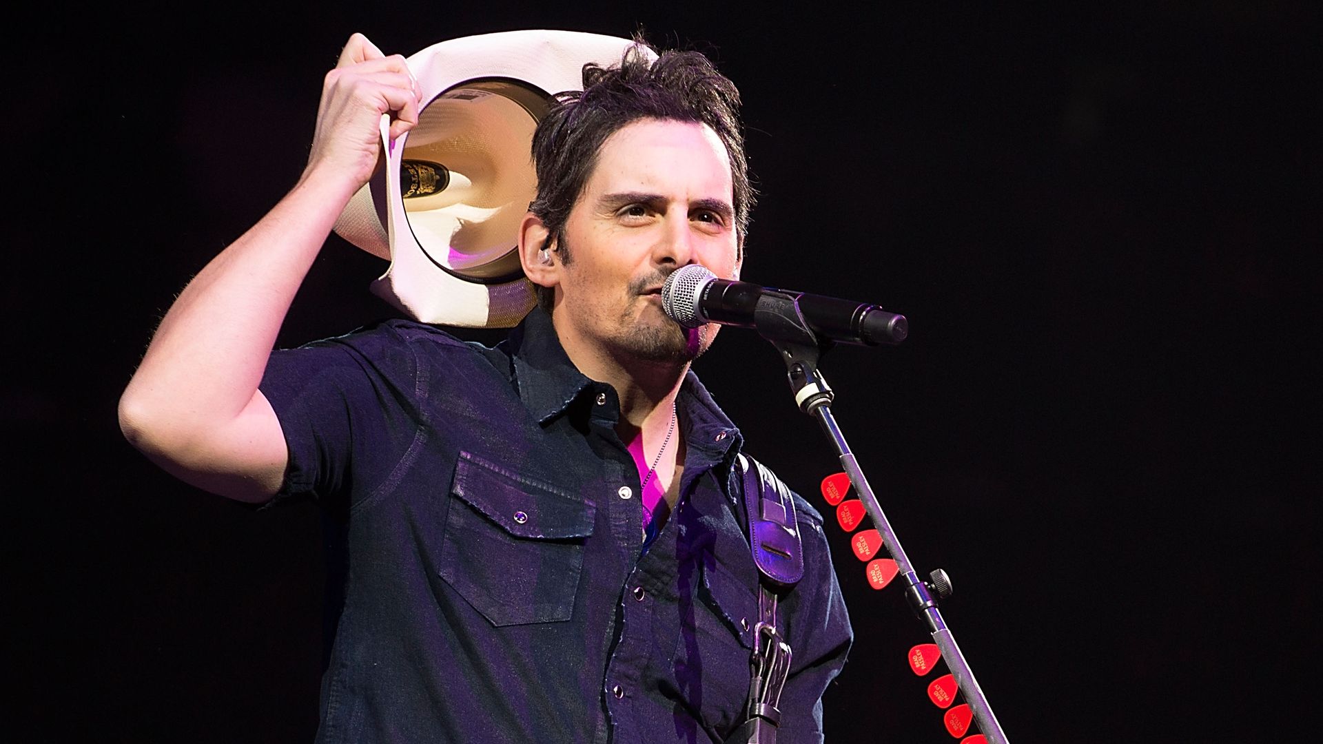 Singer songwriter Brad Paisley, wearing a black short sleeve button-up shirt, removes his cowboy hat as he sings into a microphone 