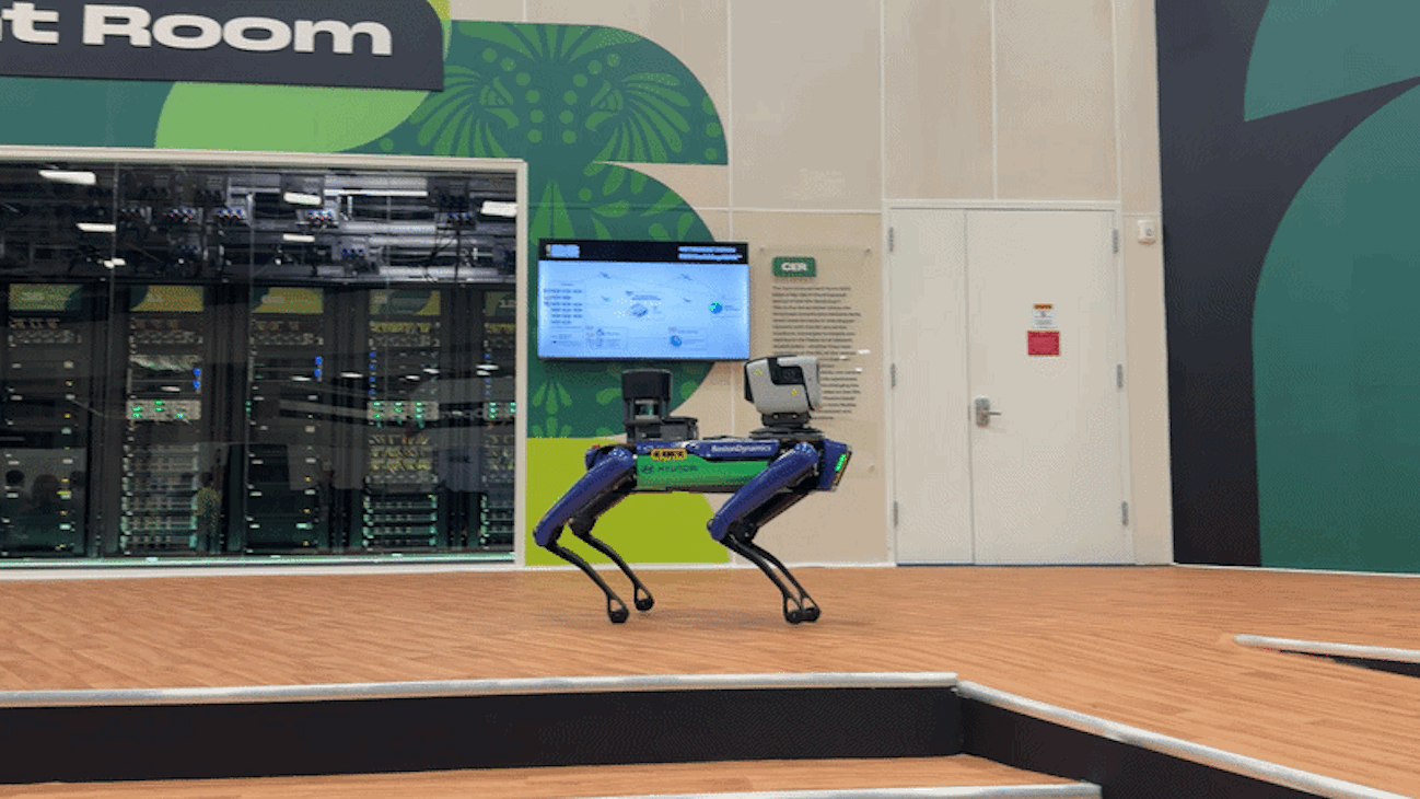 A blue four-legged robot dog with a mounted camera on its back stands on a wooden arena floor inside a tech lab. Behind it, glass-walled server racks, a wall mural, a monitor, and a white door.