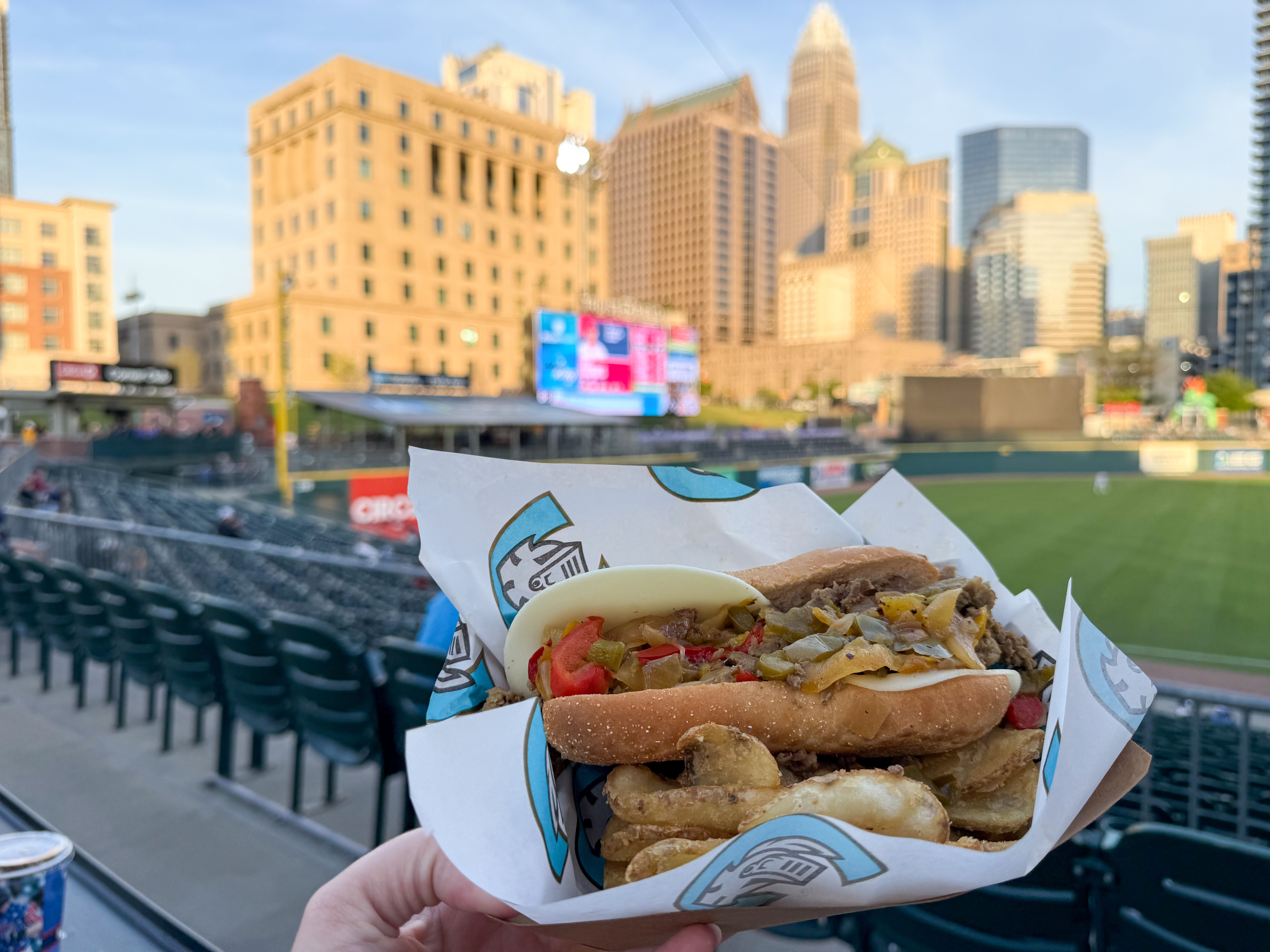 Close-up of a hand holding a loaded sandwich with peppers and onions in a bun, wrapped in branded paper, at a baseball stadium with a city skyline background.