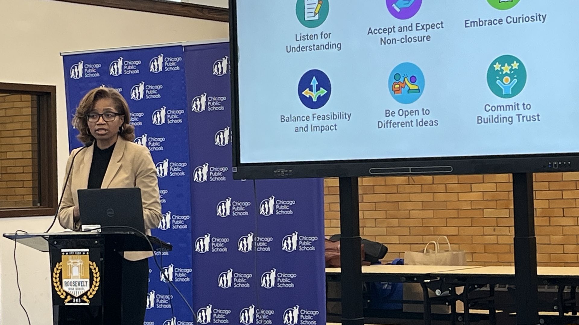 Woman in tan blazer speaking at a podium with Chicago Public Schools banners, presenting a screen showing FY2026 budget update and community norms like "Listen for Understanding" and "Embrace Curiosity".