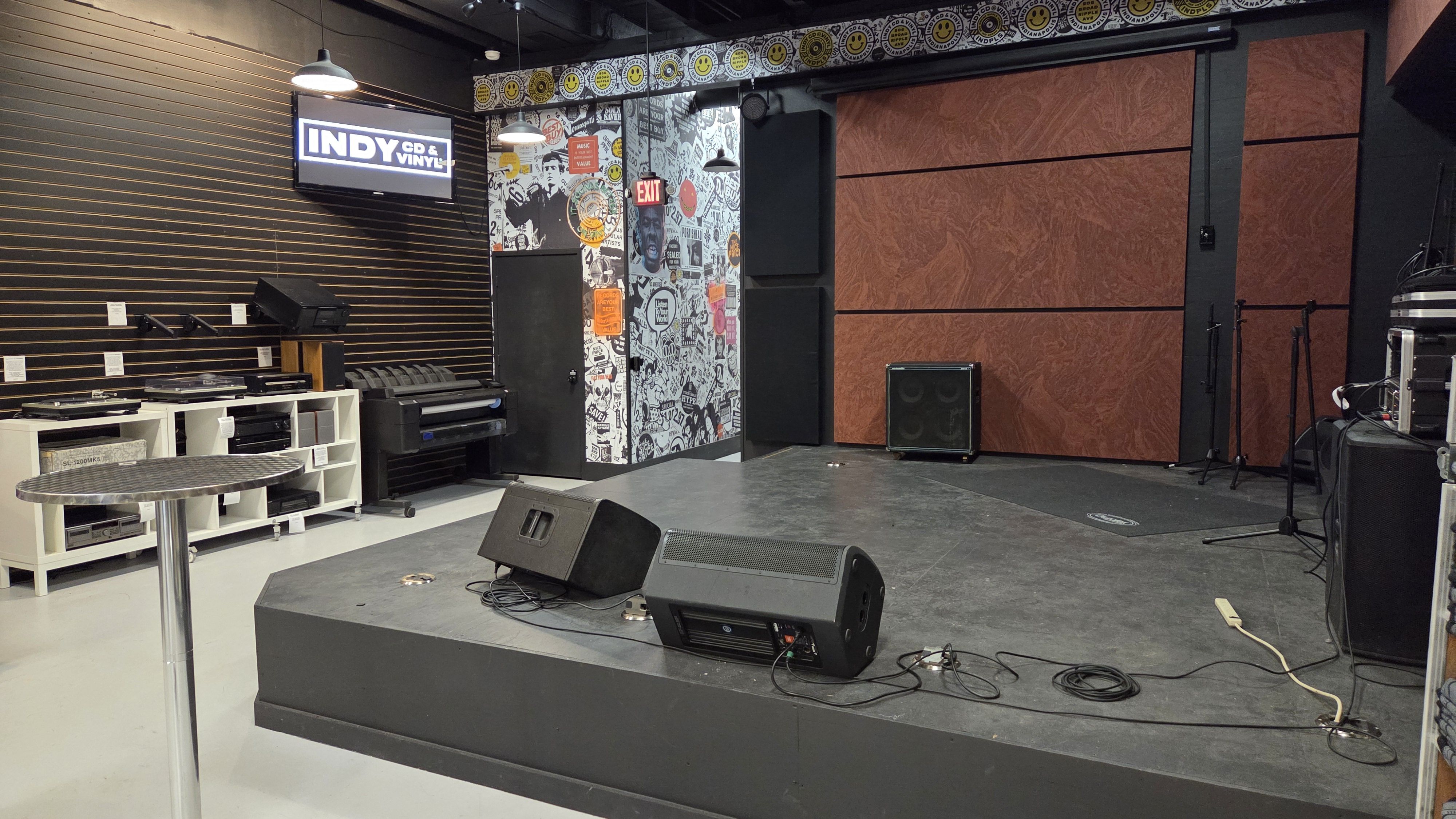 Empty small black stage with speakers, microphones, and cables in a room with black walls, a patterned wall with stickers, and a sign reading "INDY CD & VINYL".