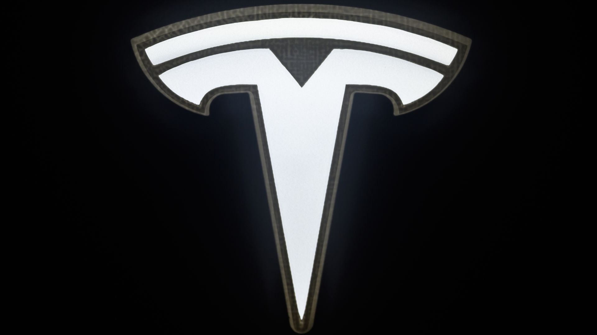 Close-up of a glowing white Tesla logo on a black background, featuring a curved top arc and a central notch that forms a stylized T.