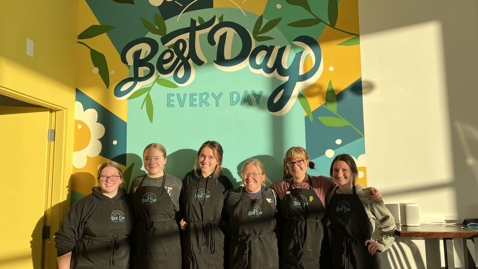 The staff of Best Day in front of a mural in the coffee shop that says "Best Day Every Day"