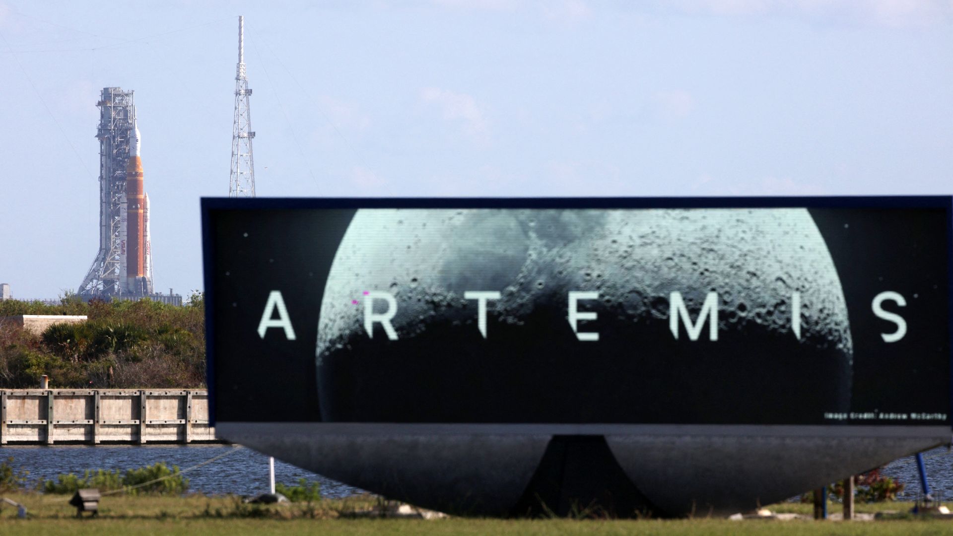 A large electronic billboard with the word "ARTEMIS" over a partial image of the moon, with a rocket on a launch pad in the distance behind it. 