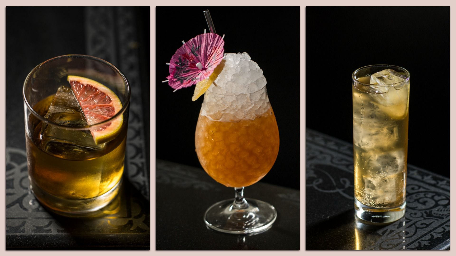 Three cocktails on a dark patterned surface: a whiskey with a large ice cube and grapefruit slice, an orange crushed-ice drink garnished with a pink paper umbrella and lemon wedge, and a tall fizzy golden drink with ice cubes.