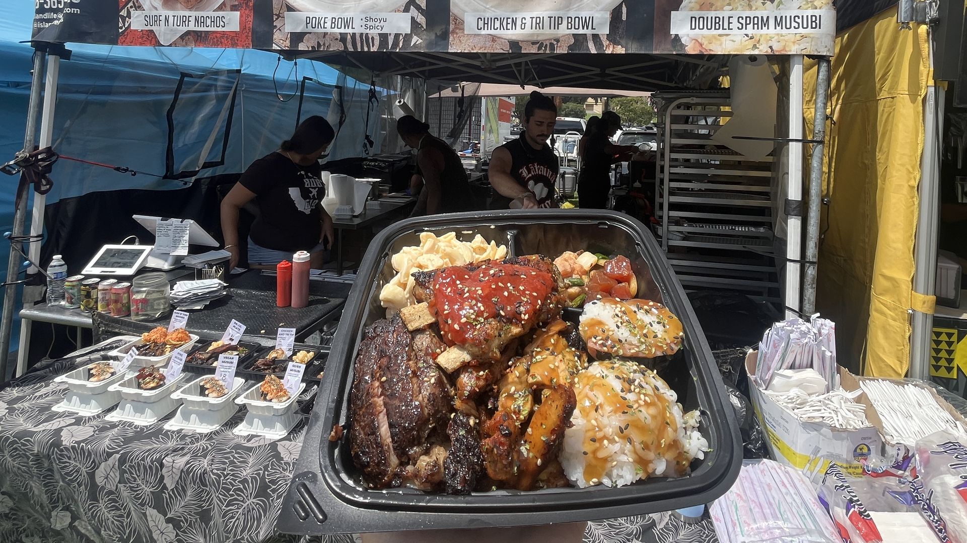Food stall named Island Life Foods at an outdoor market with a large sign showing menu items like Surf N Turf Nachos, Poke Bowl, Chicken & Tri Tip Bowl, and Double Spam Musubi. A person holds a tray of assorted grilled meats and rice.