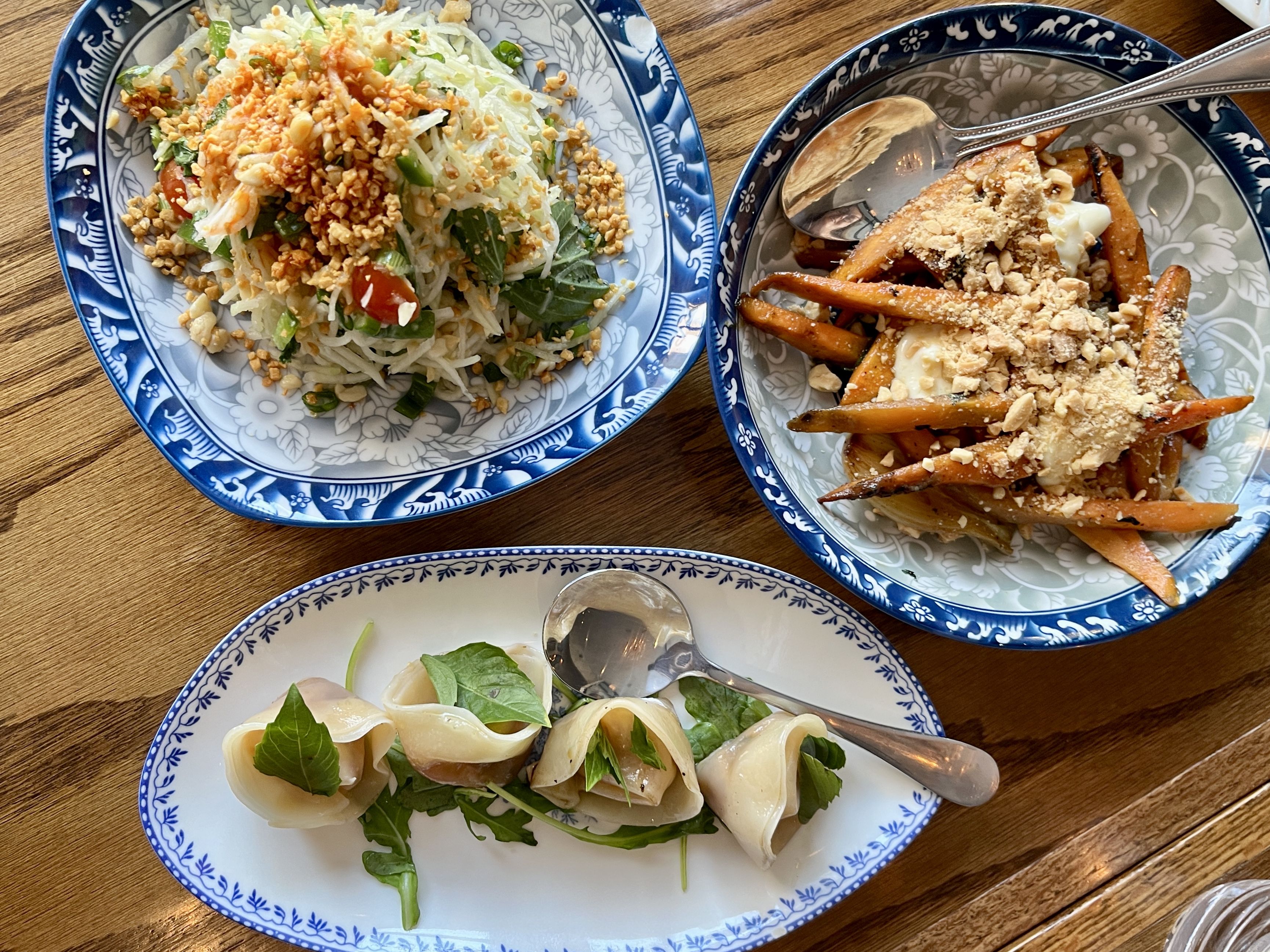 Three dishes on wooden table: shredded salad with peanuts and herbs on blue patterned plate, roasted carrot sticks with sauce and nuts on similar plate, and four dumplings with greens on white blue-rimmed plate.