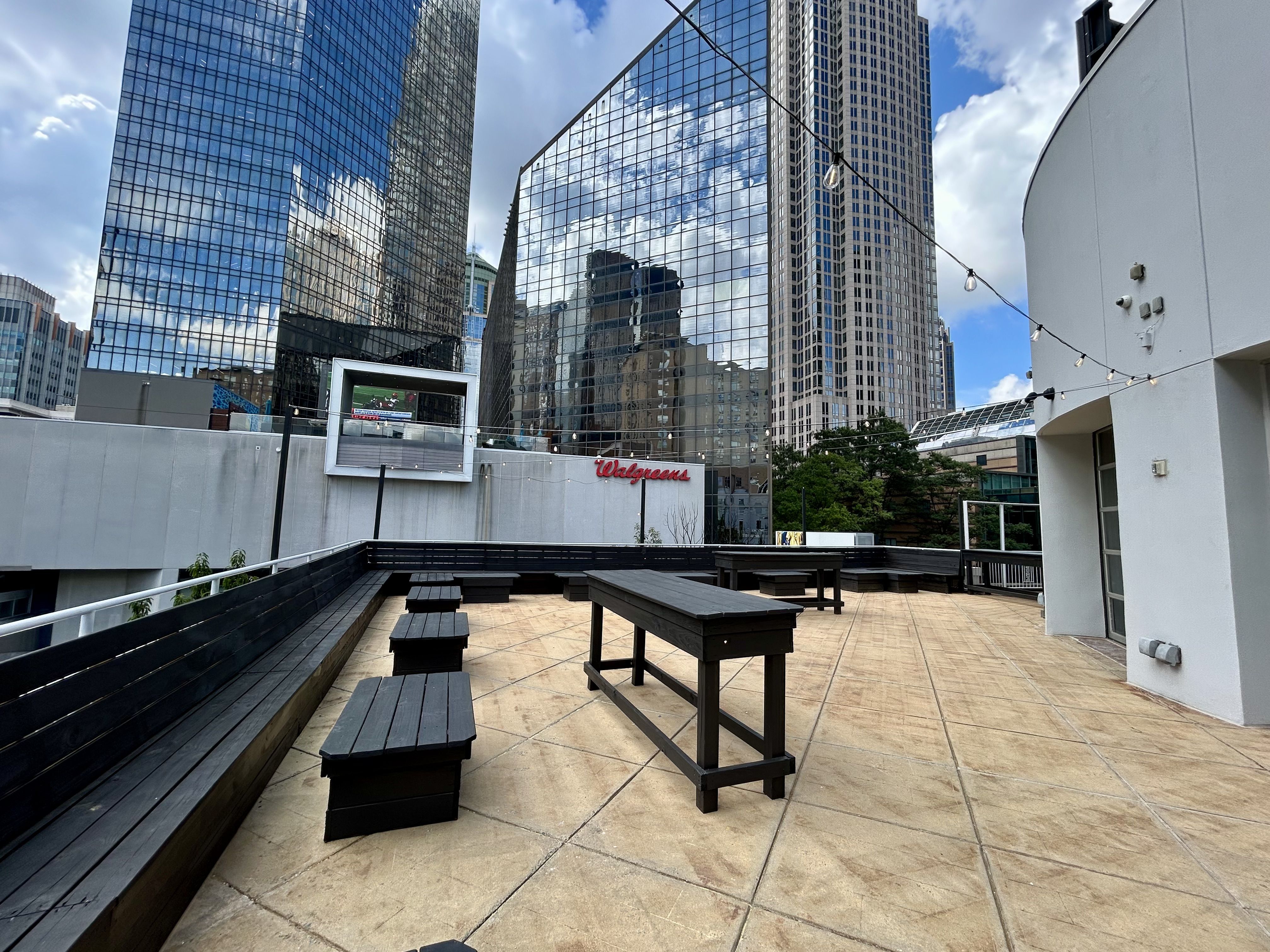 Circa Charlotte club opening uptown. Rooftop terrace with long black wooden benches and tables on a beige tiled floor, string lights overhead. Glass skyscrapers reflect blue sky and clouds; a Walgreens sign is visible.