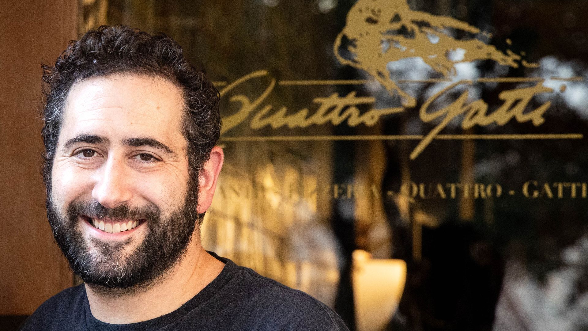 Partial portrait of a smiling man with dark hair and beard, wearing a dark T-shirt, cropped to the left of the frame, in front of a glass door with a gold logo and blurred interior.