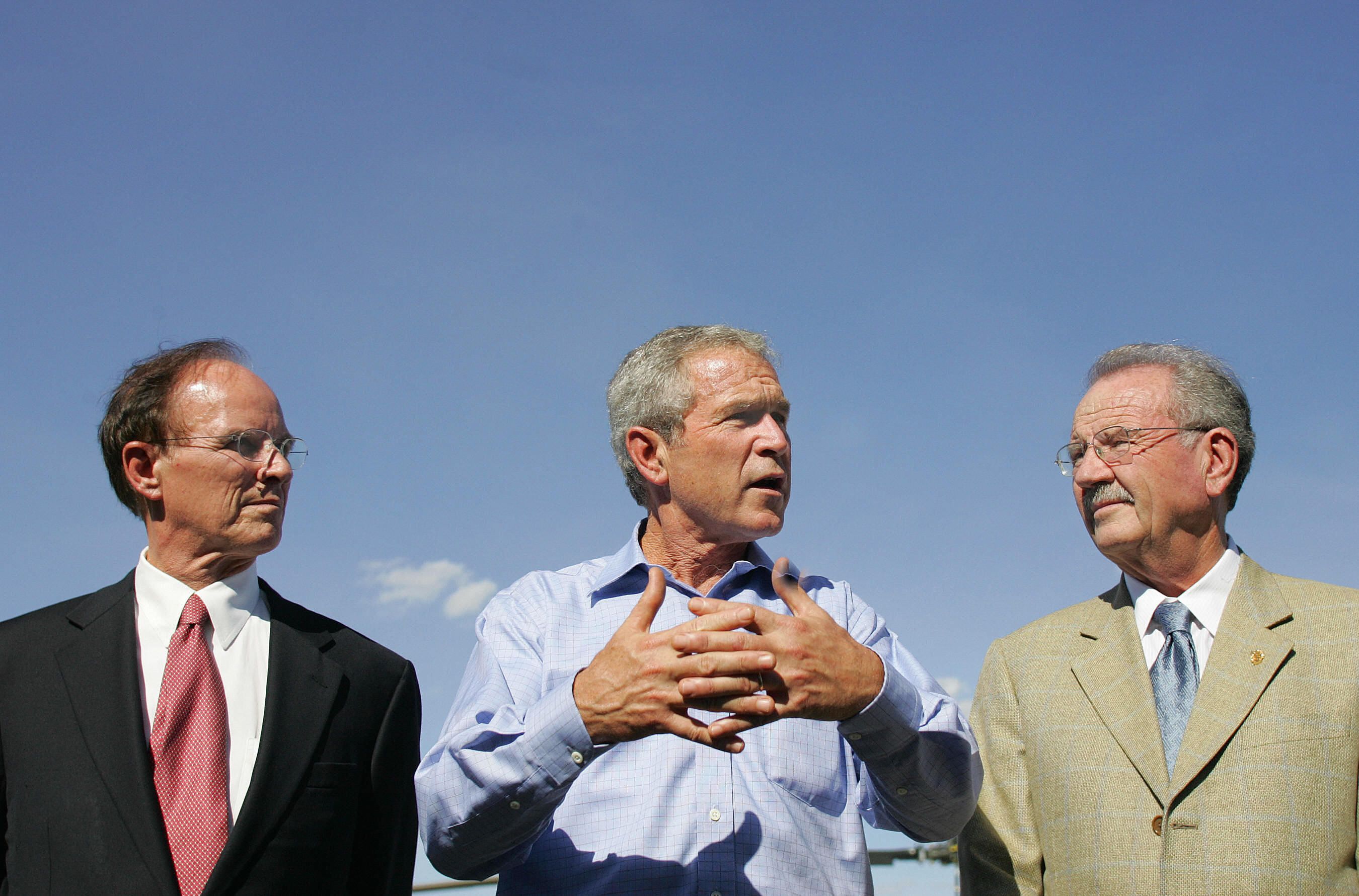 Three men stand outdoors against a clear blue sky; the man in the center speaks with hands gesturing, wearing a light blue shirt, the man on left wears a dark suit with a red tie, and the man on the right wears a tan suit with a blue tie.