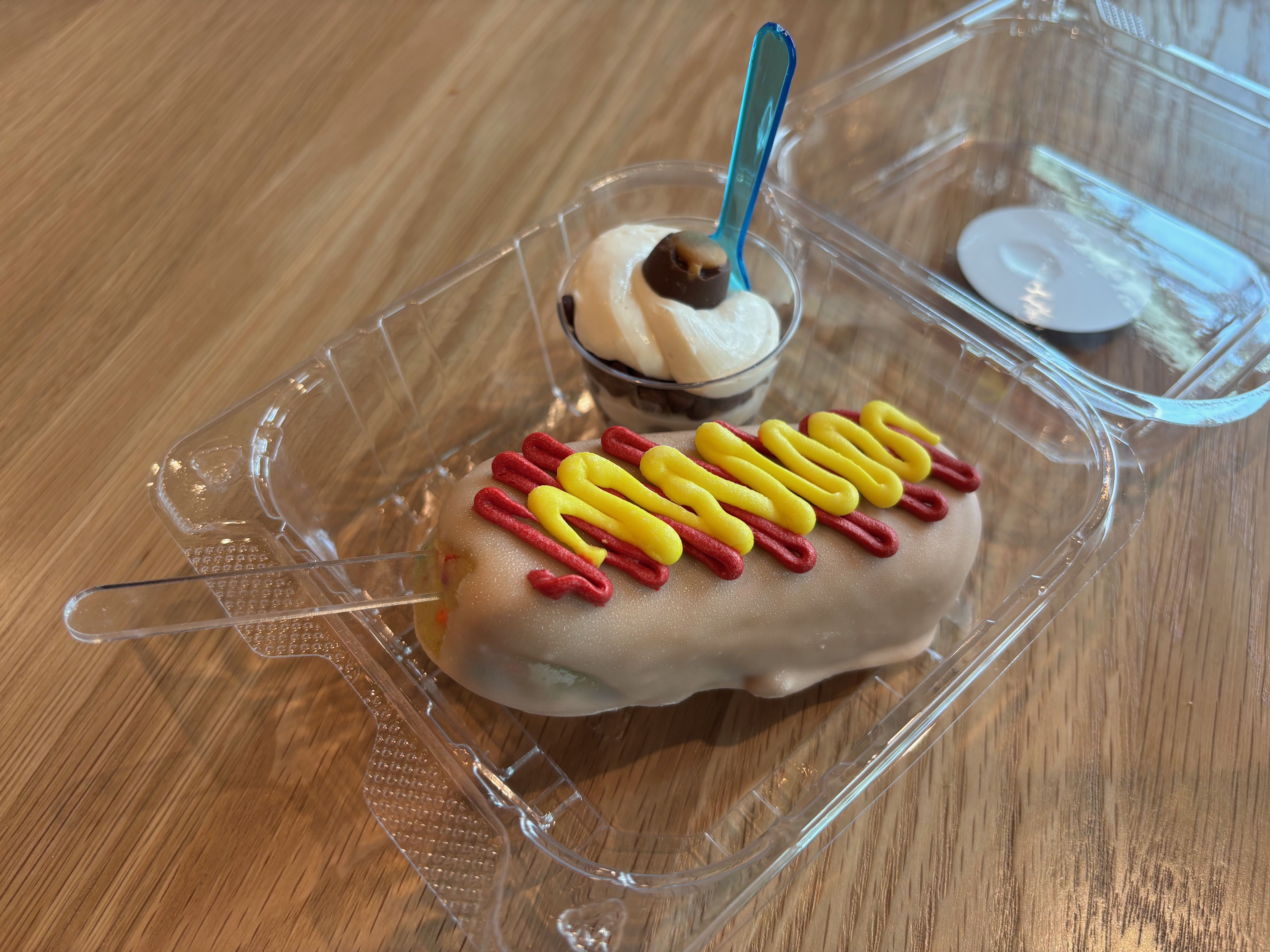 Close-up of a glazed donut inside a container, decorated with red and yellow icing swirls. A small cup holds whipped cream cheese with a Buckeye on top; a blue spoon stands upright beside it on a wooden table.