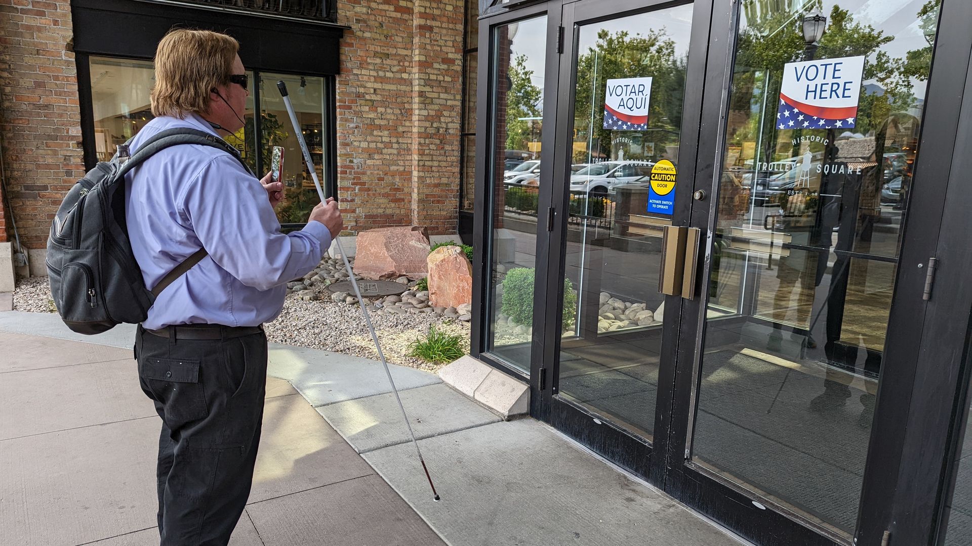 A man holds a probing cane and a phone camera up to a door with a sign reading "Vote here."