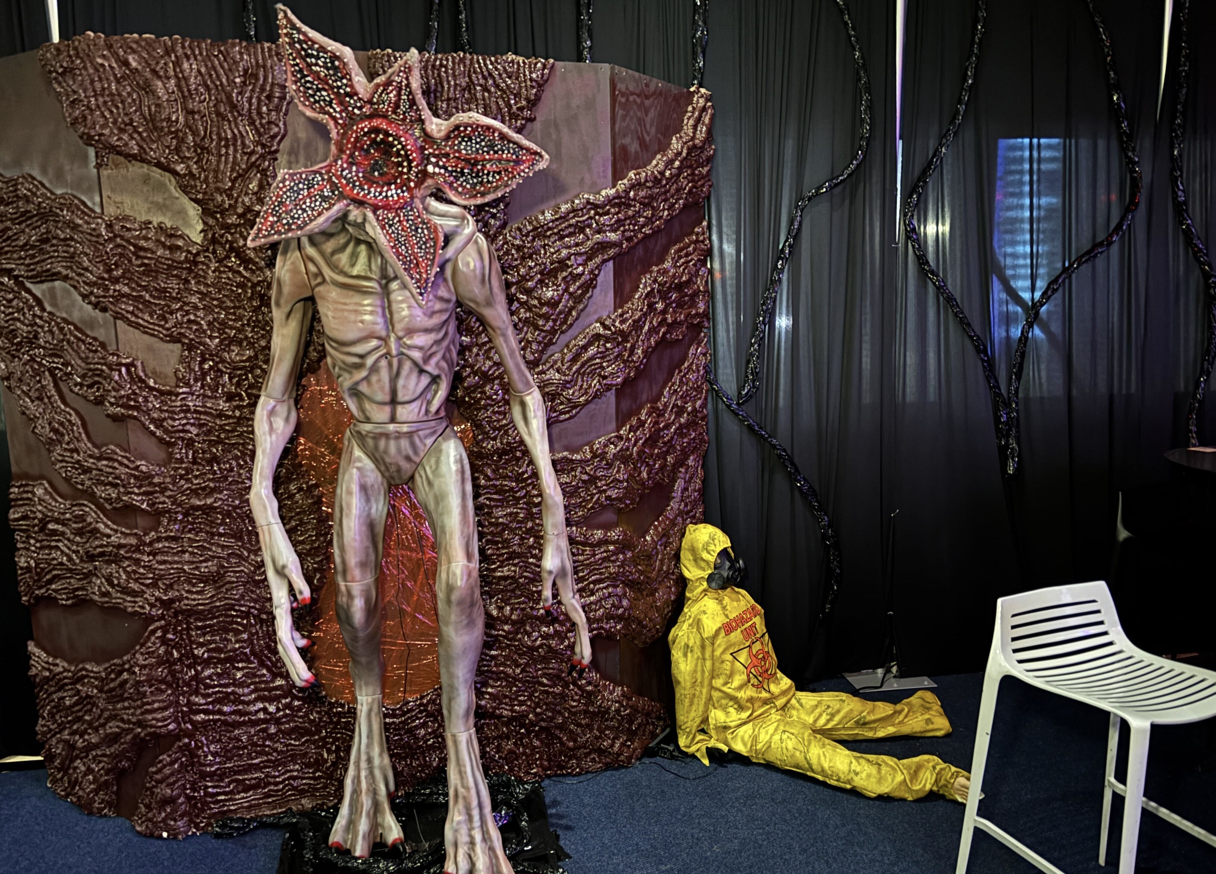 Life-size model of a creature with a starfish-like head and long limbs against a textured brown wall, next to a mannequin in a yellow hazmat suit sitting on the floor under black drapes.
