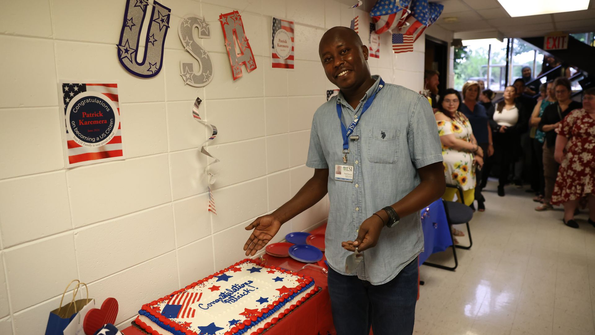 A smiling man in a light denim shirt with a blue lanyard stands beside a red, white, and blue flag-themed cake on a table, as patriotic USA decorations and a crowd line the hallway.