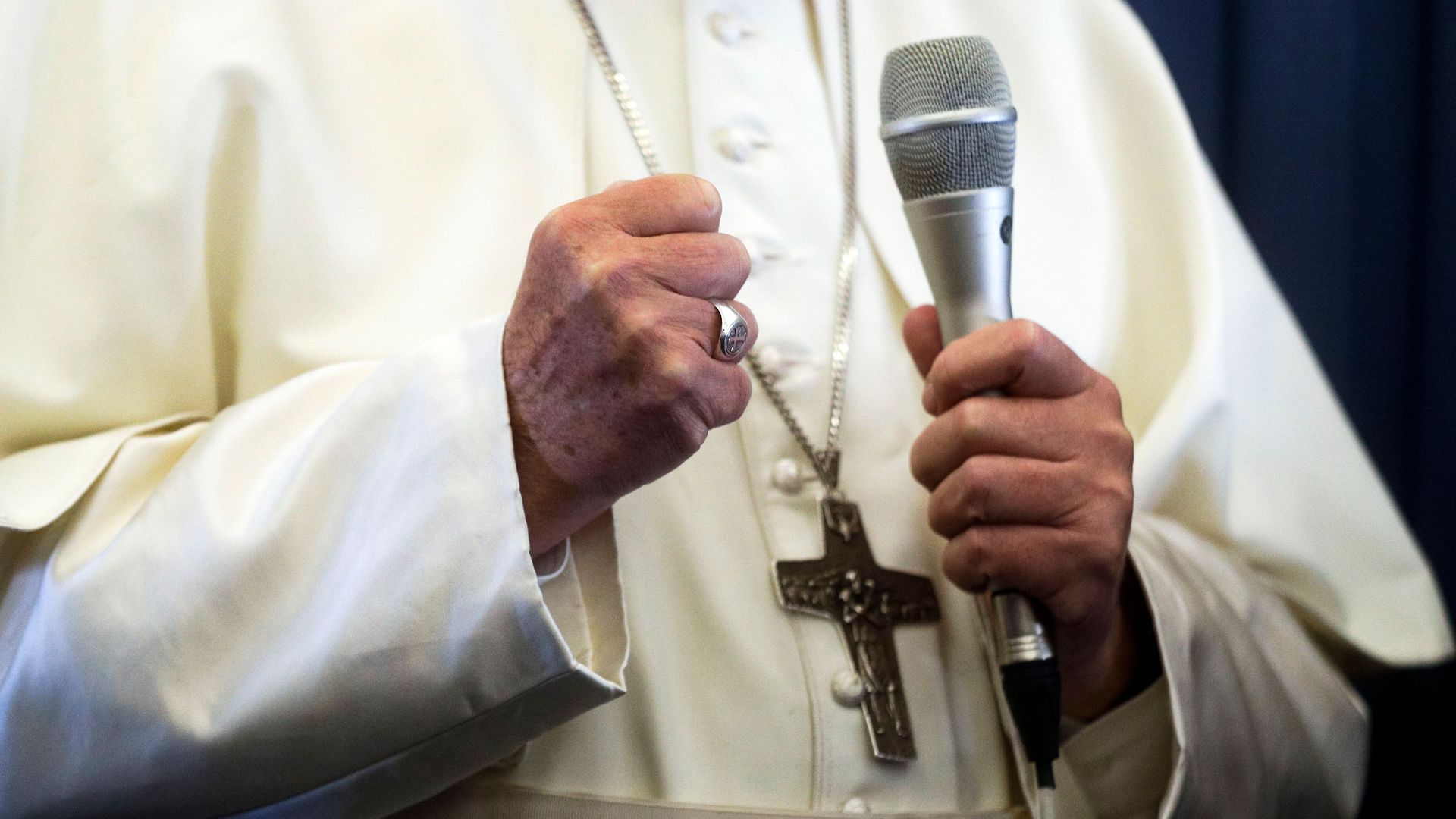 A shot of the pope's hands and robes, but not his face. One hand is in a fist, one hand holds a microphone. A crucifix necklace hangs on his white robes. 