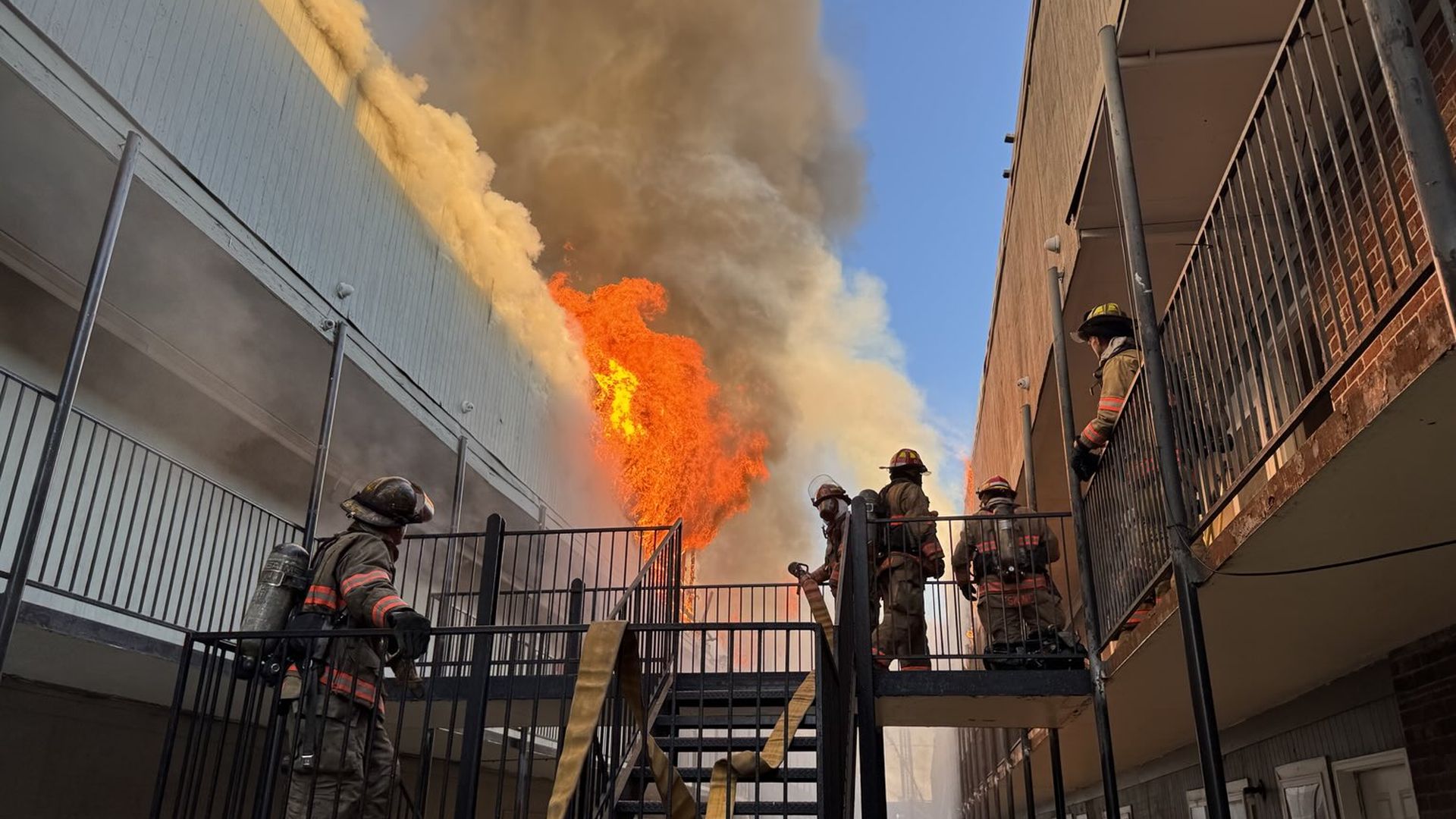 Firefighters in full gear stand on black metal stairs and balcony, battling a large orange fire and thick gray smoke between two multi-story buildings under a clear blue sky.