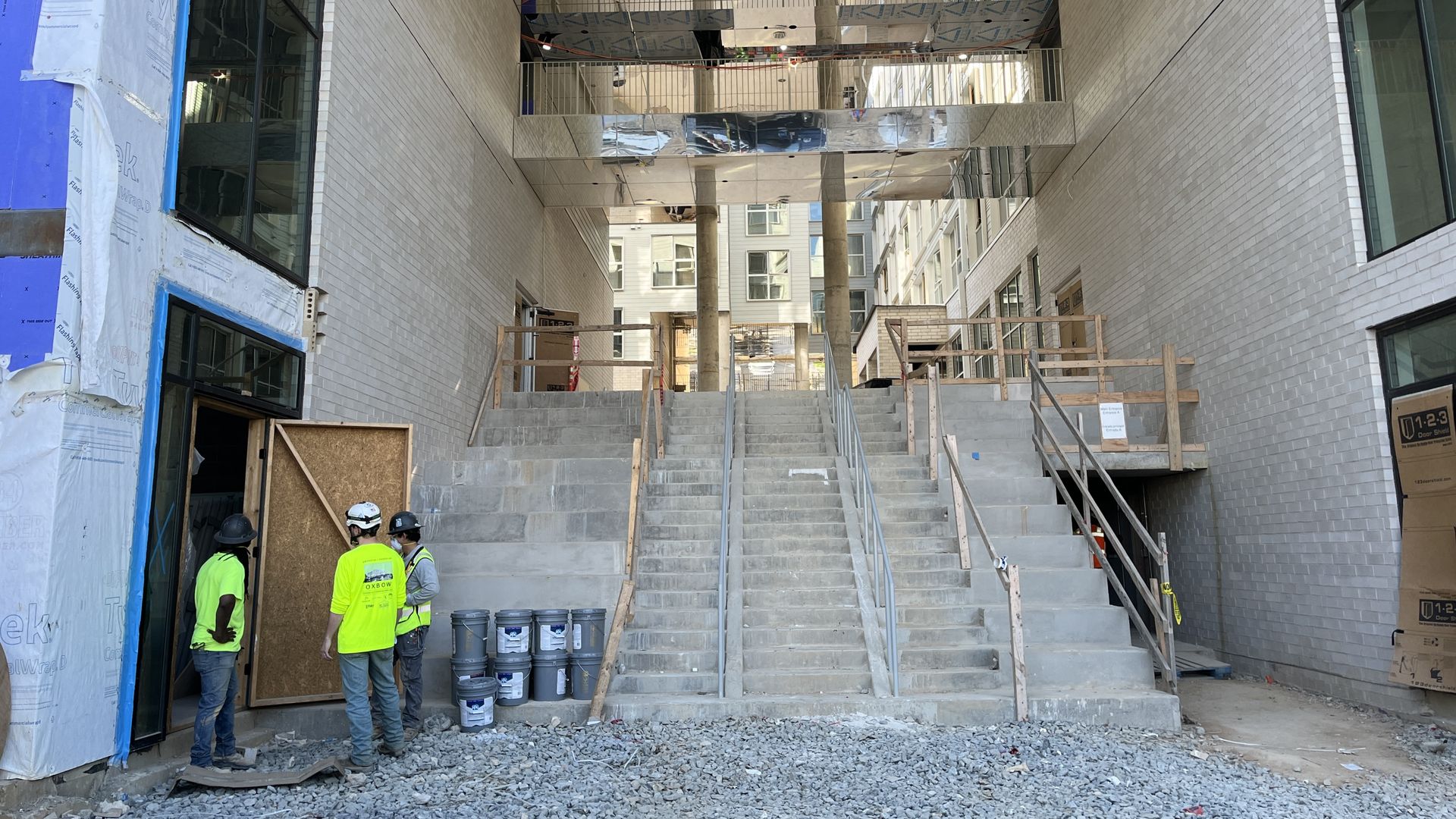 Construction site in a modern white-brick building. Three workers in neon vests and hard hats stand by concrete stairs; blue wrap on left, reflective metal panels overhead, gravel and buckets nearby.