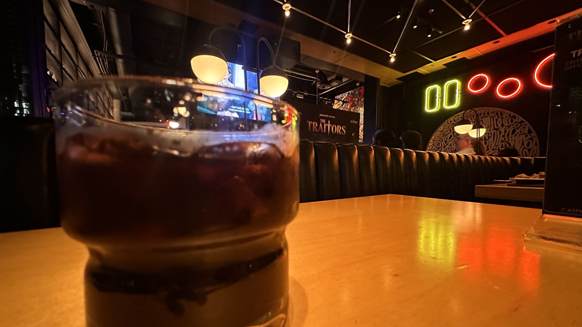 Close-up of a dark drink with ice on a wooden table in a dimly lit bar or restaurant with black leather seating, neon lights, and a TV screen showing "The Traitors".