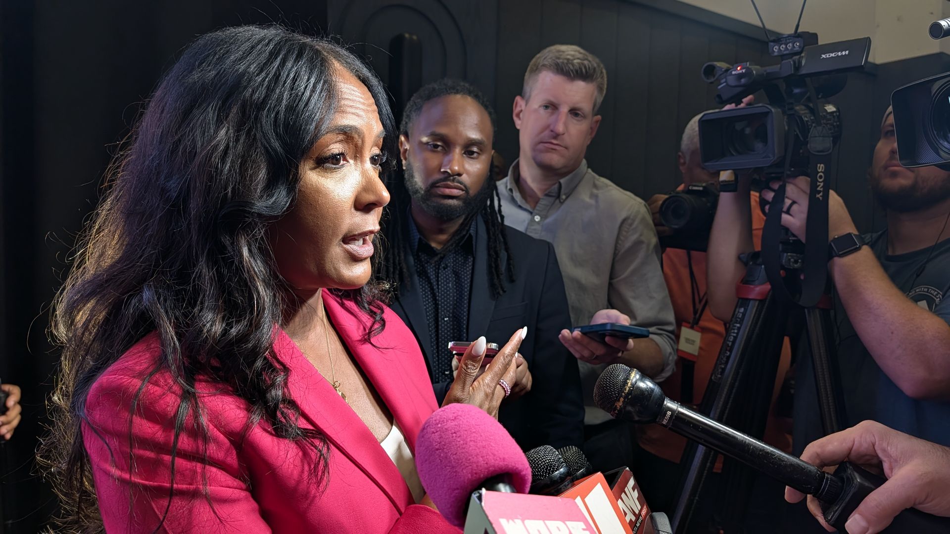A woman in a bright pink blazer speaks to reporters, surrounded by microphones with pink foam and several journalists filming.