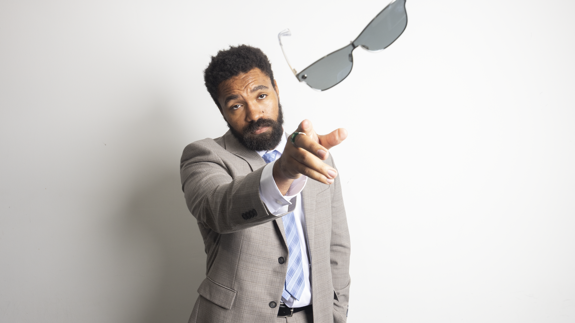 Bearded man in a beige suit and blue striped tie points at the camera against a pale background, with gray sunglasses floating above him.