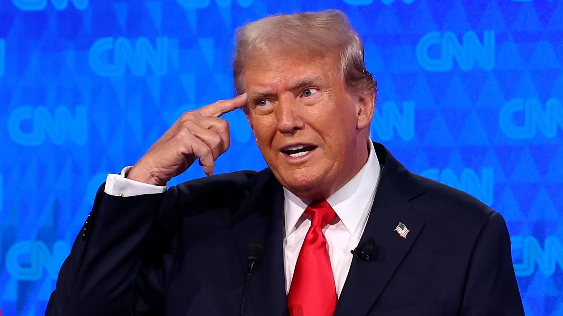Donald Trump, in a blue suit, white shirt and red tie, points to his right temple with his index finger.