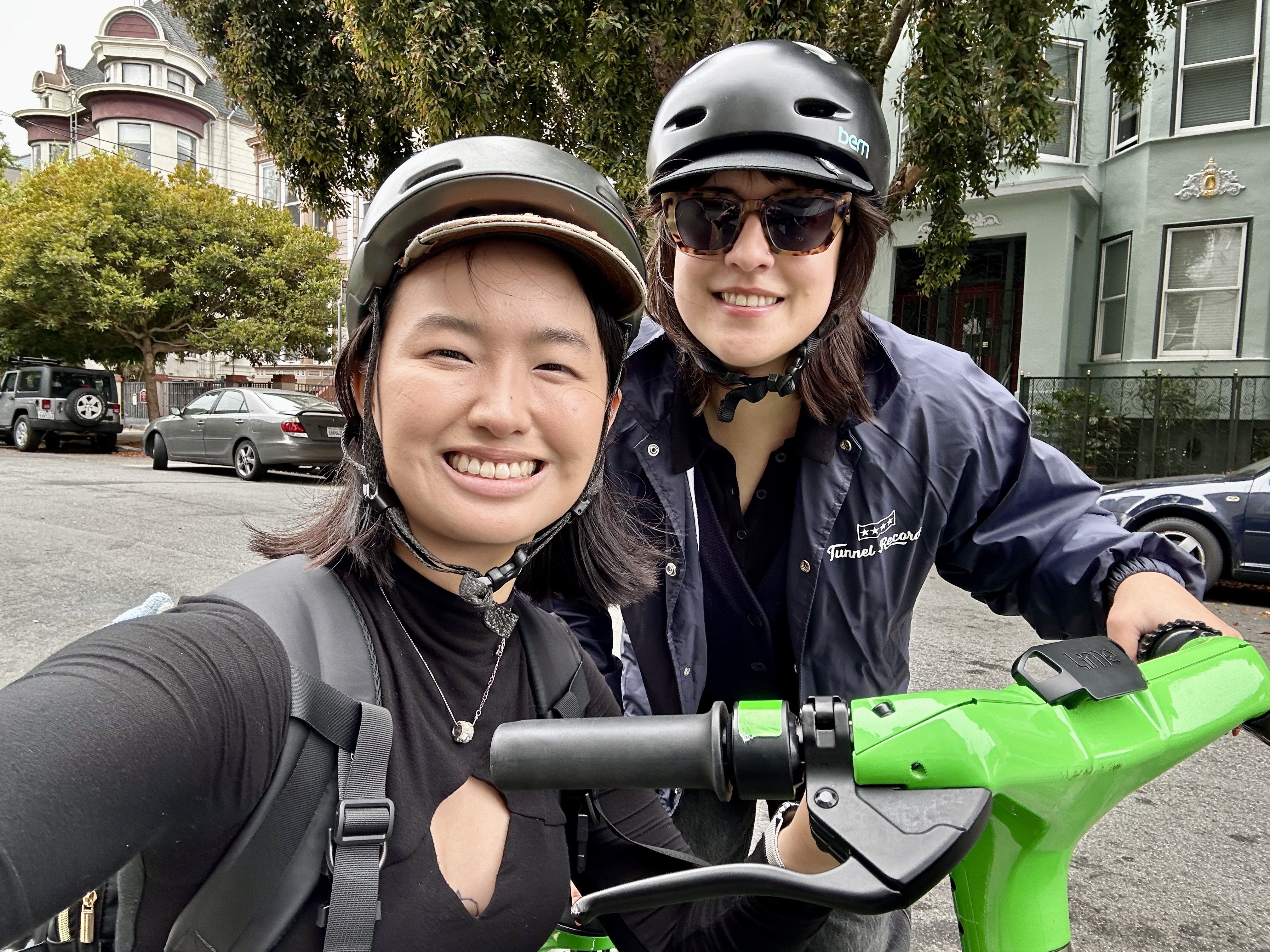 Two smiling people wearing black helmets, one in a black shirt and the other in a navy jacket, standing with a bright green Lime electric scooter on a city street with houses in the background.