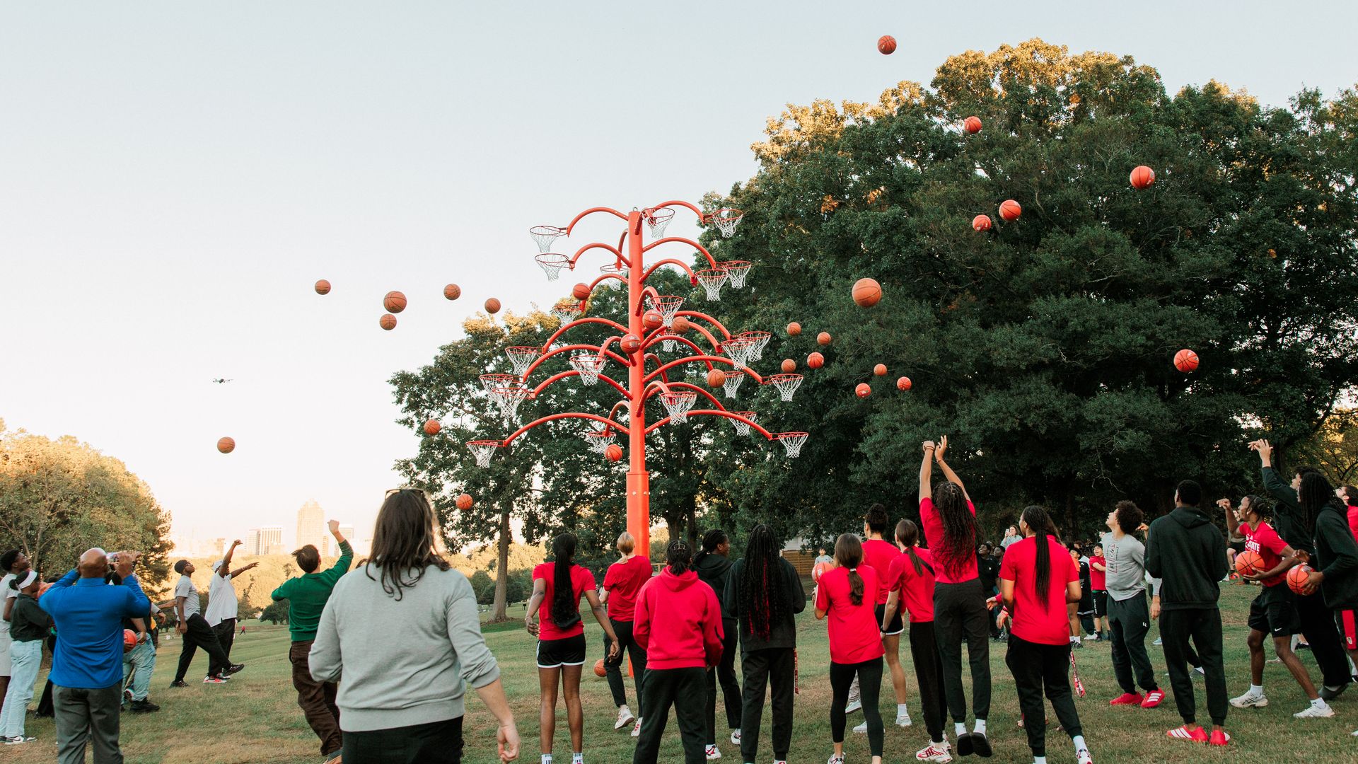 Group of people in a park, many in red shirts, watch a tall red multi-hoop basketball sculpture as basketballs fly through the air near a large tree against a pale blue sky.