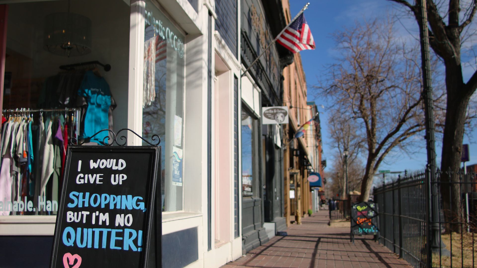 Black sidewalk sign in front of a clothing store reads, "I WOULD GIVE UP SHOPPING, BUT I'M NO QUITTER!" with colorful letters. American flag and bare trees line the sunny Colorado Avenue, running through Old Colorado City in Colorado Springs.
