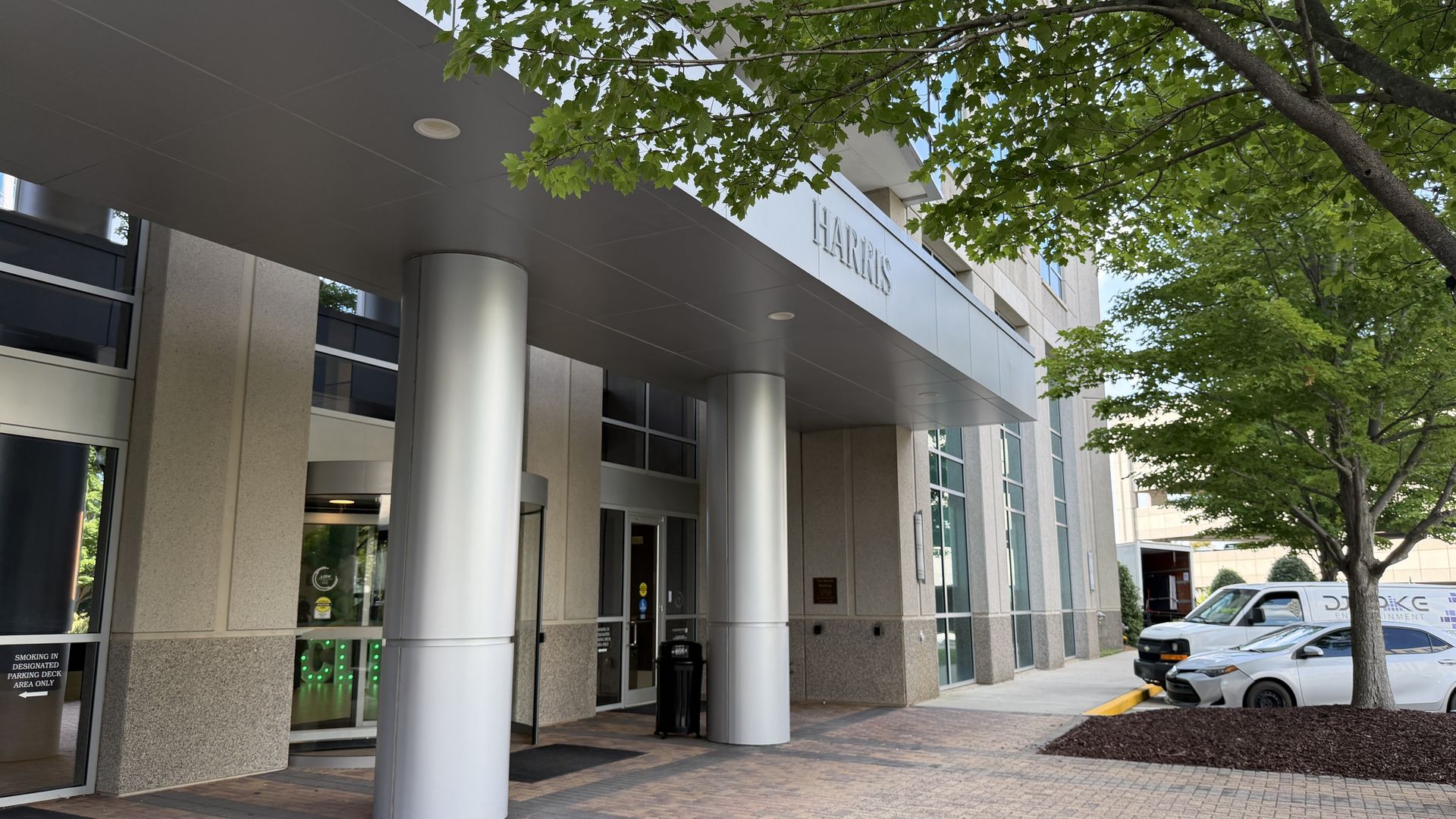 Modern building entrance with a grey canopy on silver columns, green leafy trees overhead, and a sign reading HARRIS on the façade; glass windows and a parked car nearby.