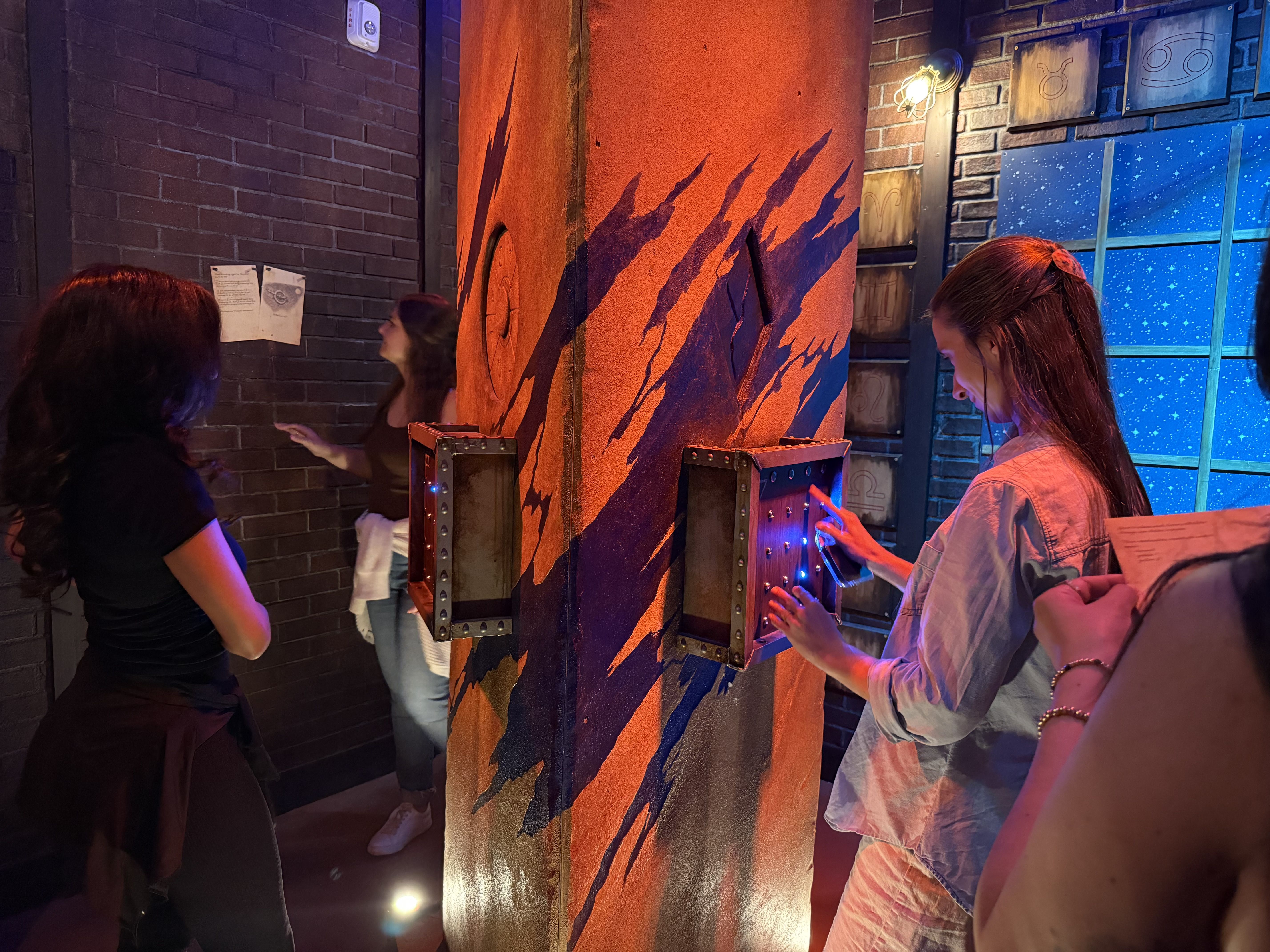 Dim arcade scene with a tall orange striped pillar. People touch blue-lit control panels on it, brick walls and starry blue panels in the background.