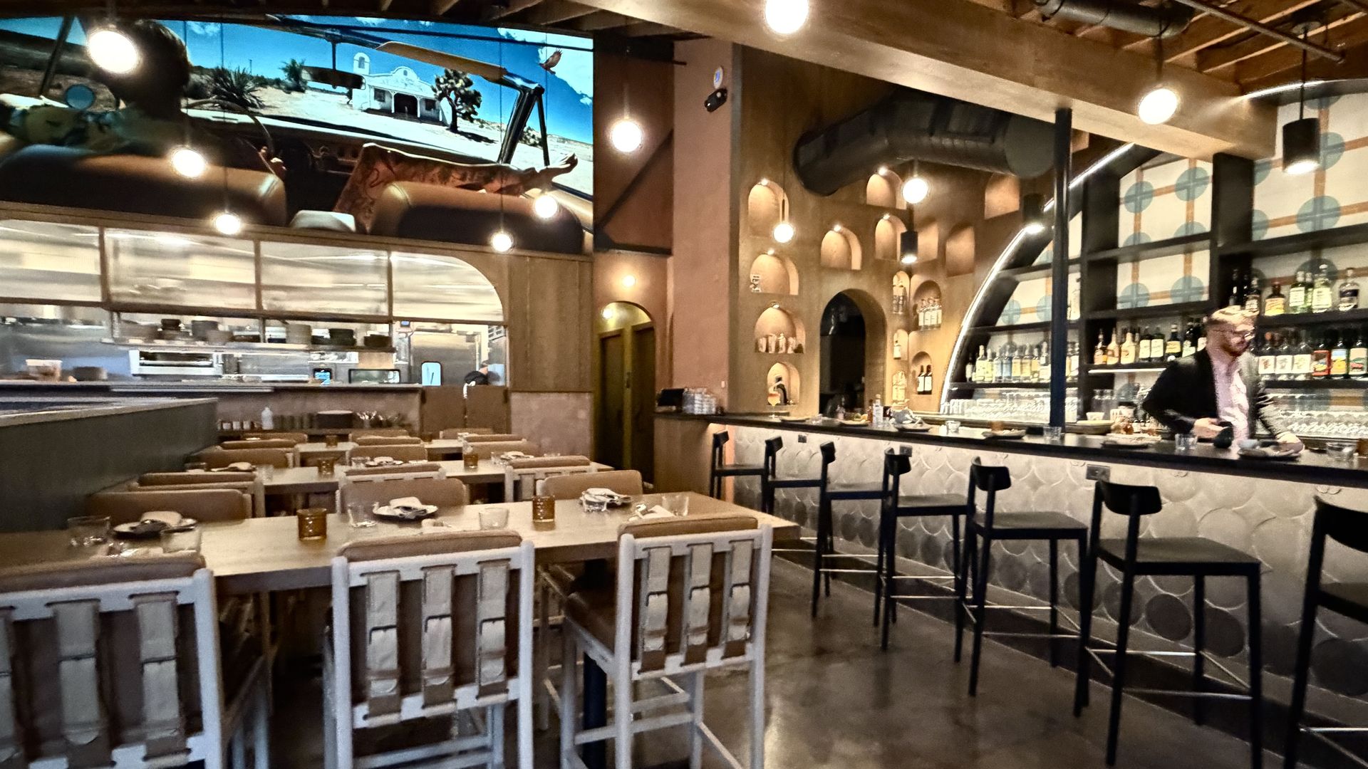 Interior of a modern restaurant with wooden tables and padded chairs on the left, a bar area with black stools on the right, warm lighting, a large screen showing a desert scene.