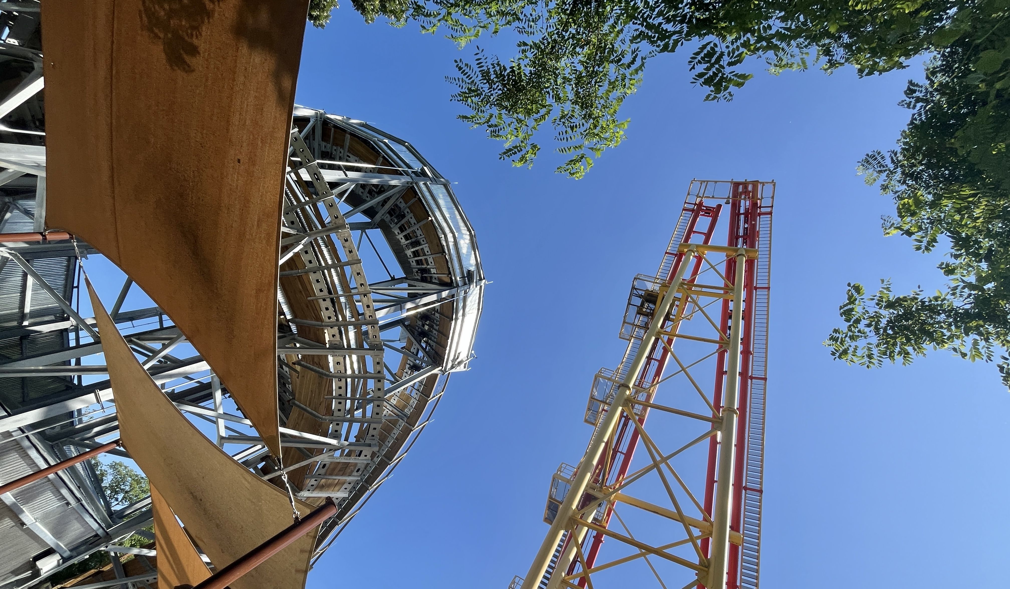 Blue sky with green trees frames a steel roller coaster featuring a circular loop on the left and a tall red-yellow track on the right.