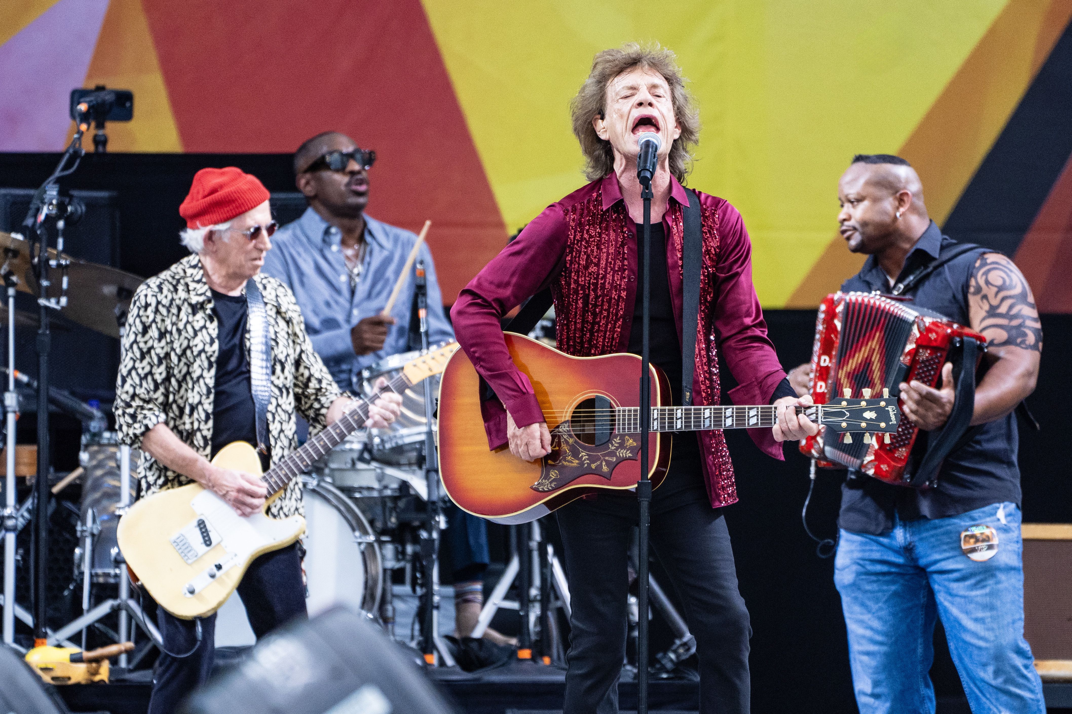 Mick Jagger sings into a microphone while holding an acoustic guitar. Dwayne Dopsie plays the accordion behind him with the rest of the Rolling Stones.