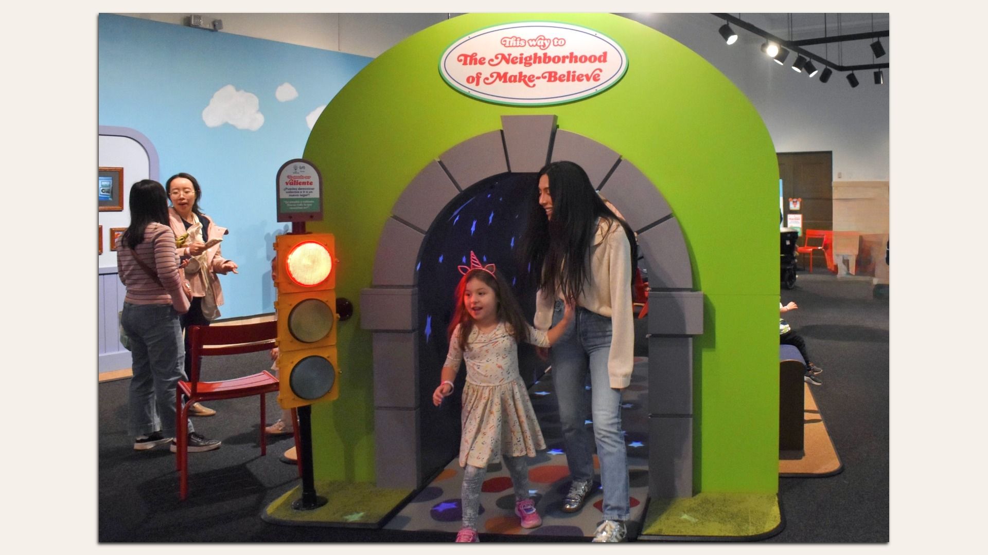 Inside a children's museum, a green archway labeled "This way to The Neighborhood of Make-Believe" frames a smiling adult guiding a young girl through a tunnel, with a traffic-light prop and blue sky mural nearby.