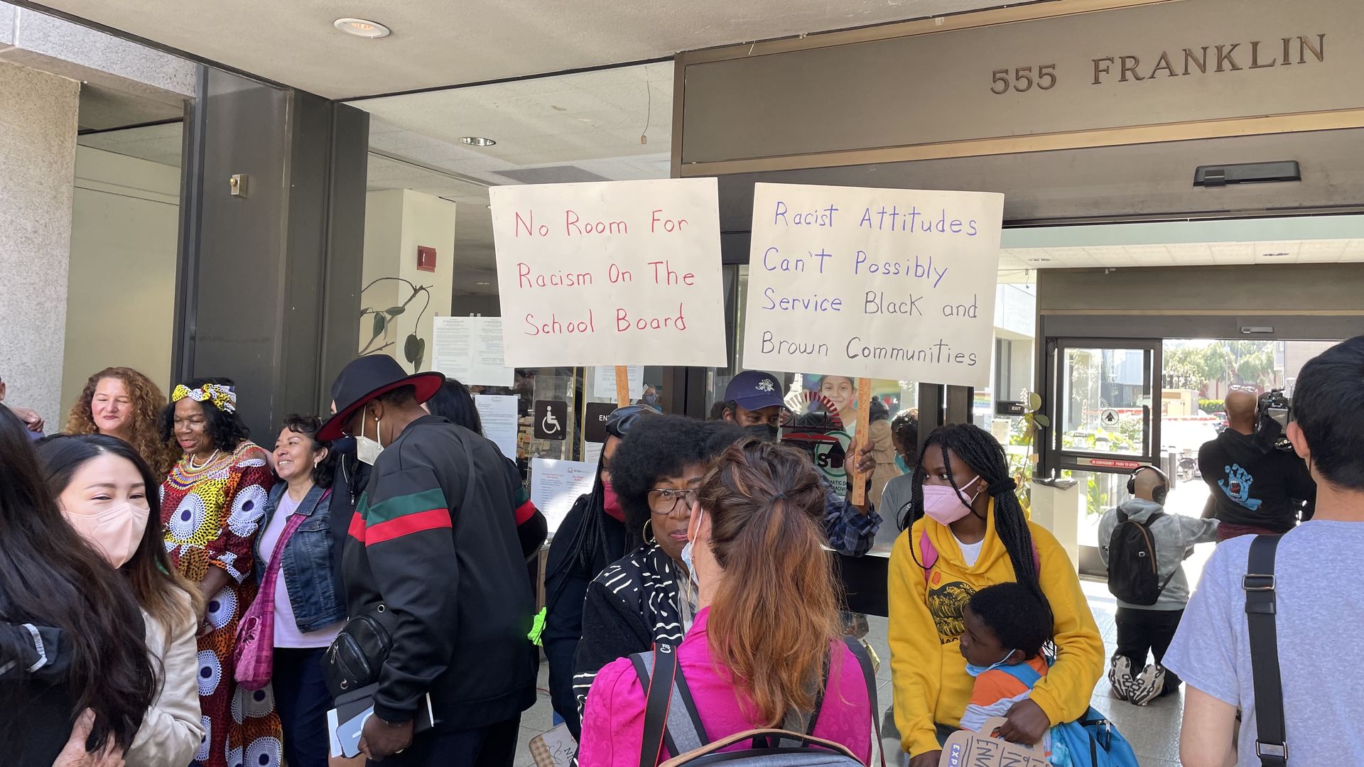 Two protest signs read "No room for racism on the school board" and "Racist attitudes can't possibly serve black and brown communities."