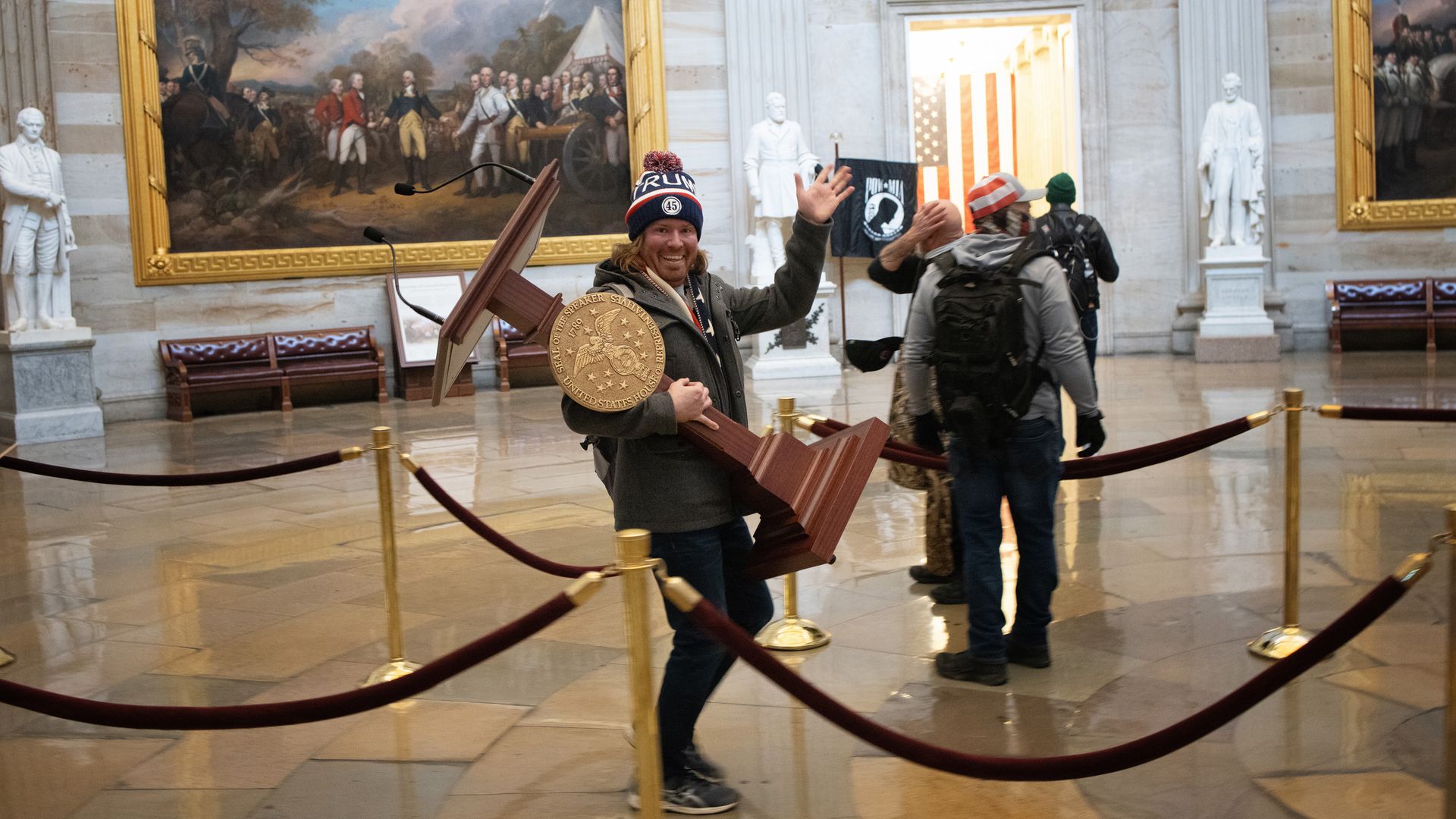 Man in beanie waves at the camera while carrying a lectern inside the capitol building.