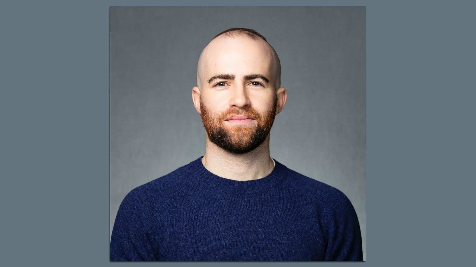 Portrait of a bald man with a reddish beard, wearing a navy blue sweater, facing the camera with a neutral expression against a gray studio backdrop.