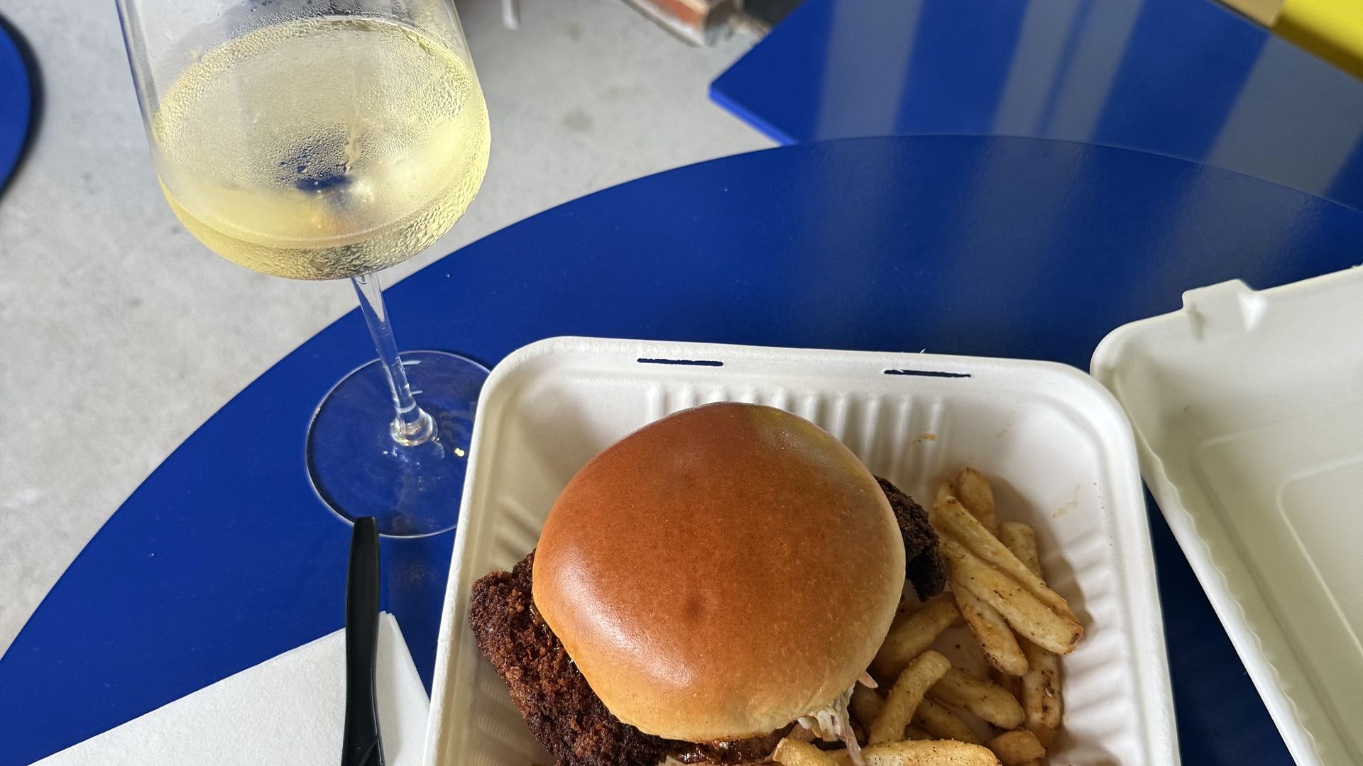 Takeout container with crispy fried chicken sandwich on a toasted bun and seasoned French fries, placed on a blue table next to a glass of chilled white wine and a black plastic knife on a white napkin.