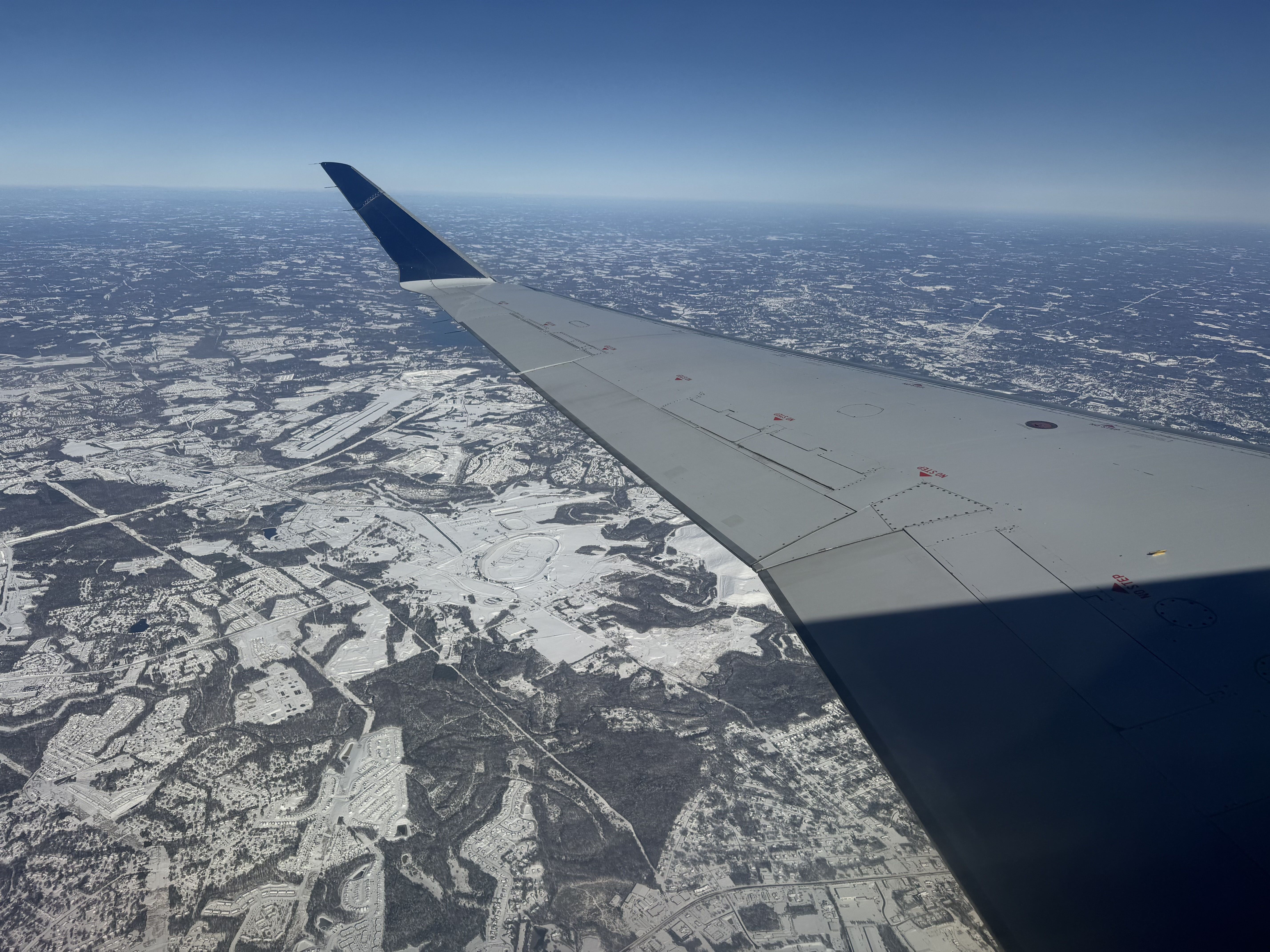 View from airplane window showing silver wing with blue tip over a snowy landscape with scattered trees and buildings under a clear blue sky.