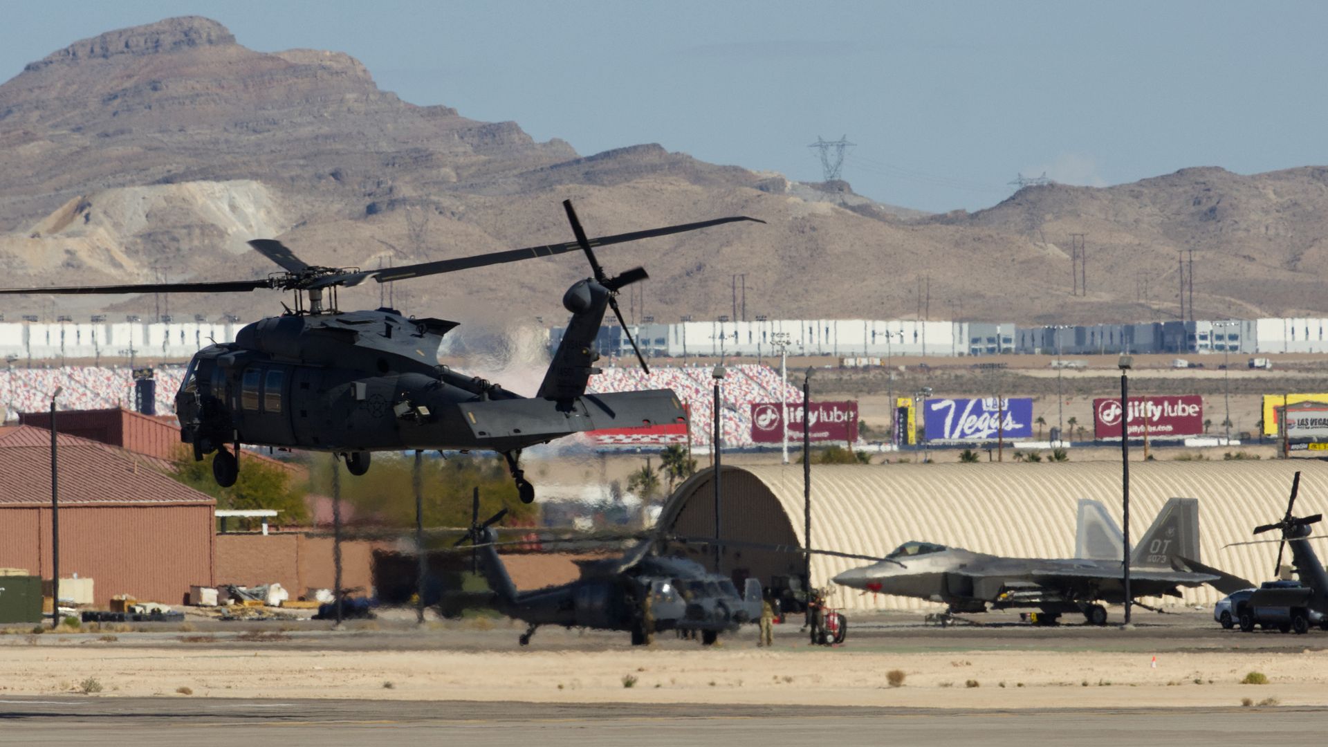 Black military helicopter hovering over desert airstrip with more helicopters and a jet near hangars. There are desert mountains in background, as well as advertisements including for Las Vegas and Jiffy Lube.