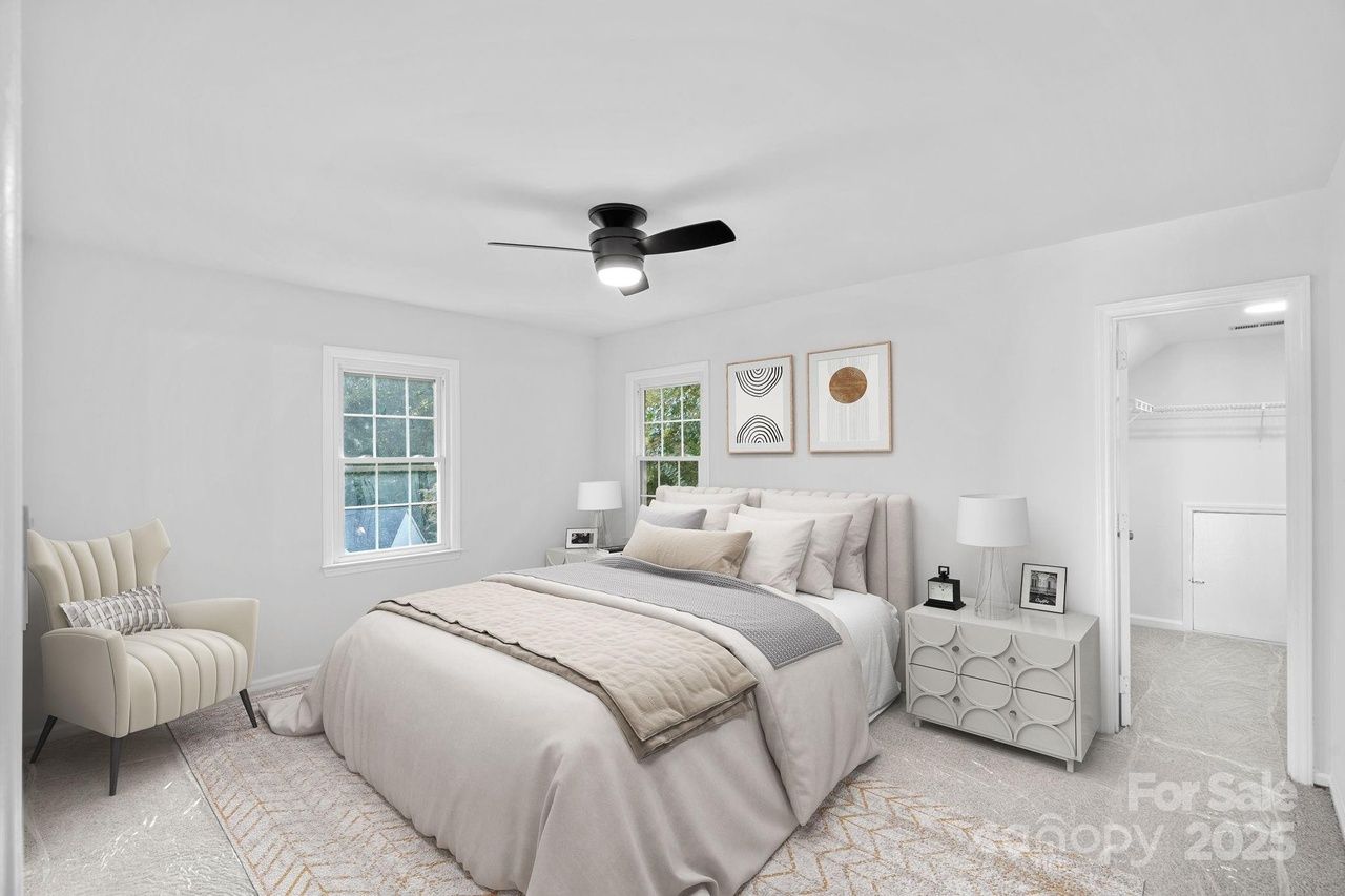 Bright bedroom with beige bed, grey and cream bedding, two abstract framed prints above the bed, cream armchair with striped pillow, white nightstands with lamps, and a ceiling fan.