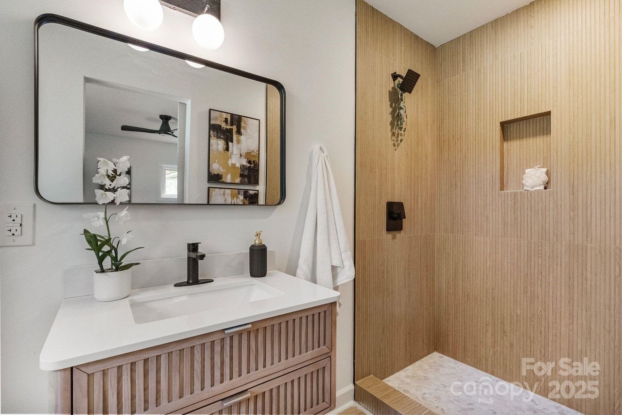 Modern bathroom with wood-textured shower walls, black shower fixtures, white countertop with black faucet, white orchid in pot, large mirror, and abstract wall art.