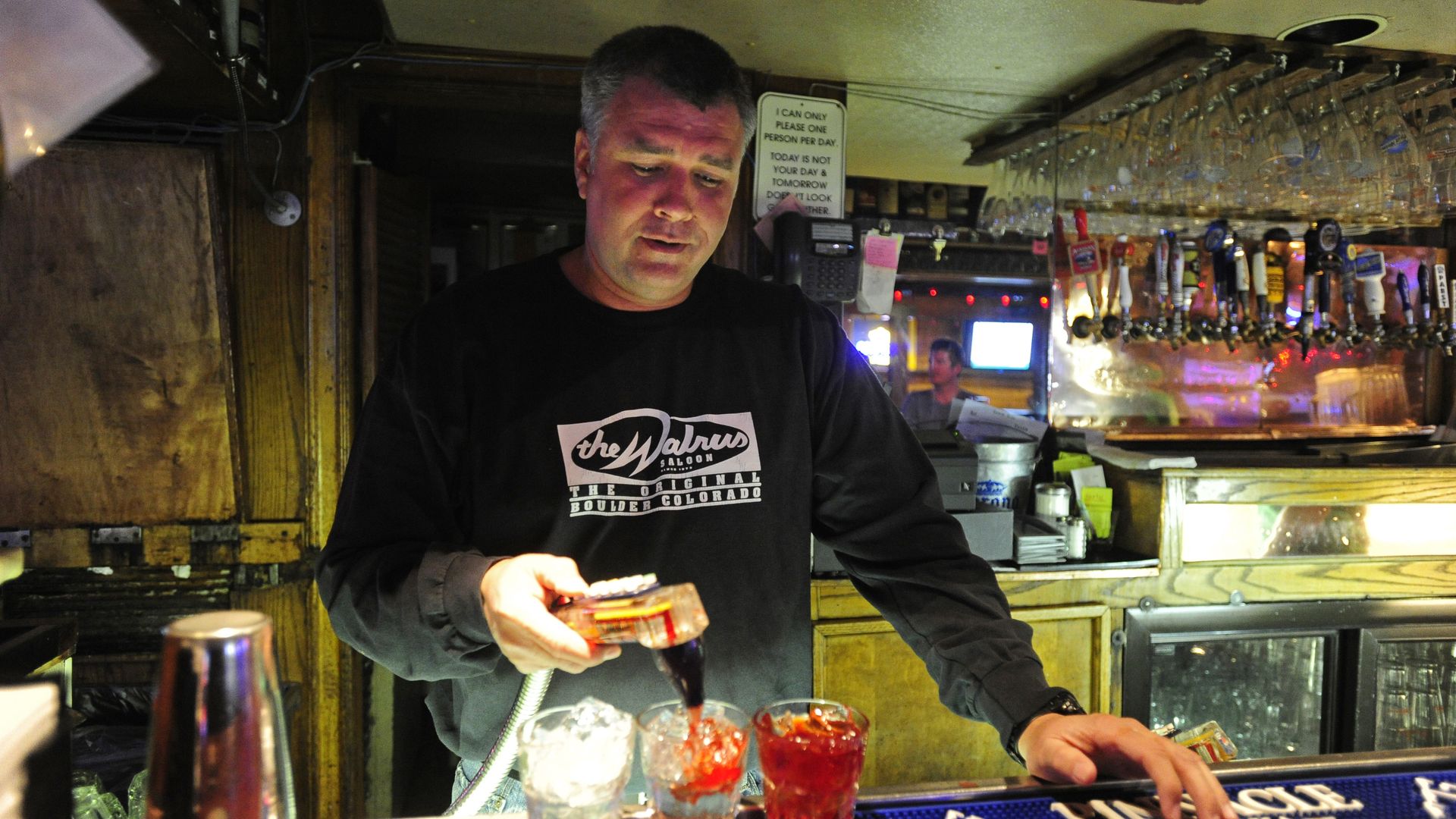 Bartender in a black "the Walrus Saloon" shirt pouring a dark liquid into a glass with ice at a wooden bar with mugs and various beer taps overhead.