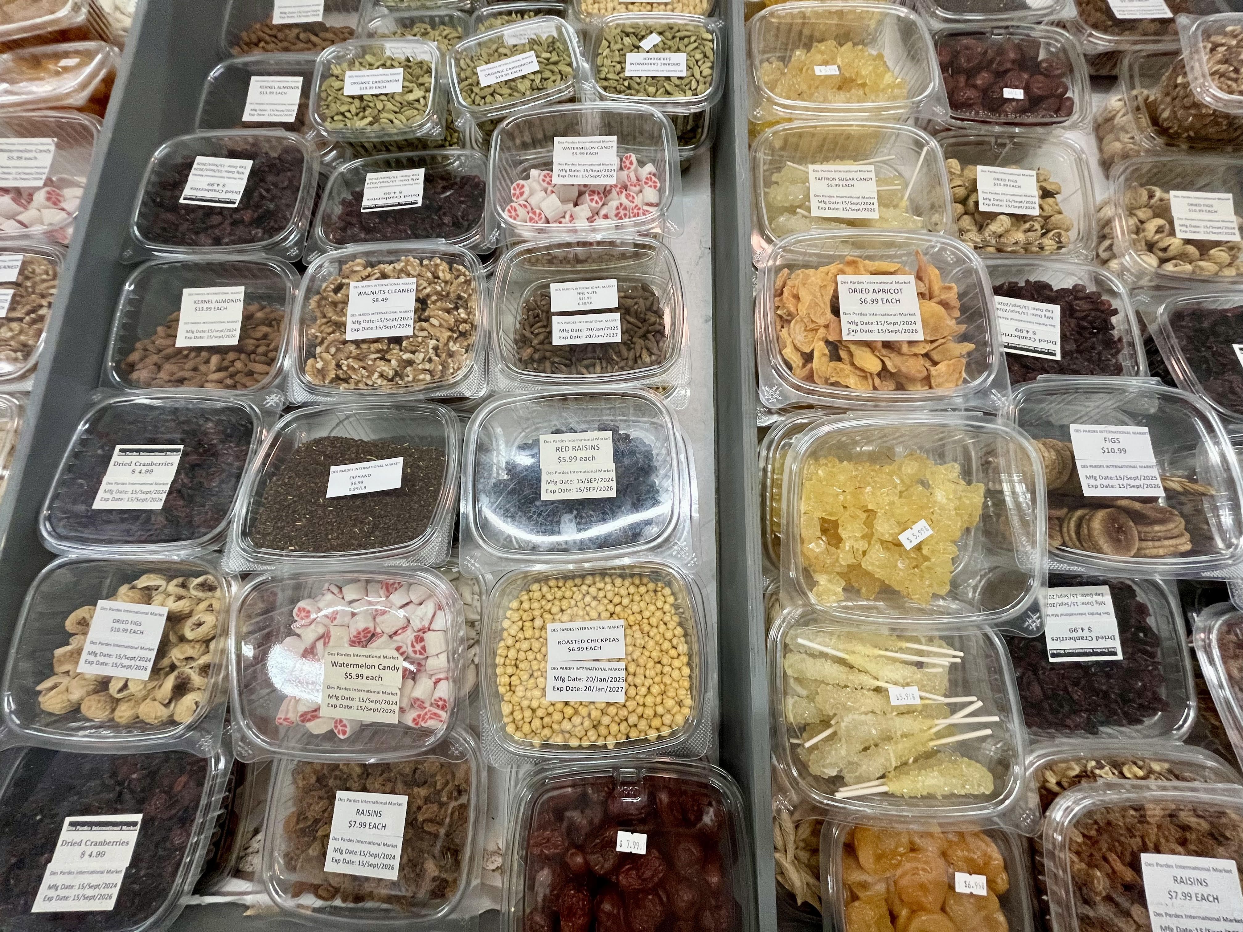 Rows of clear containers filled with assorted nuts, dried fruits, candies, and spices at Des Pardes International Market in San Antonio, labeled with prices and product names.