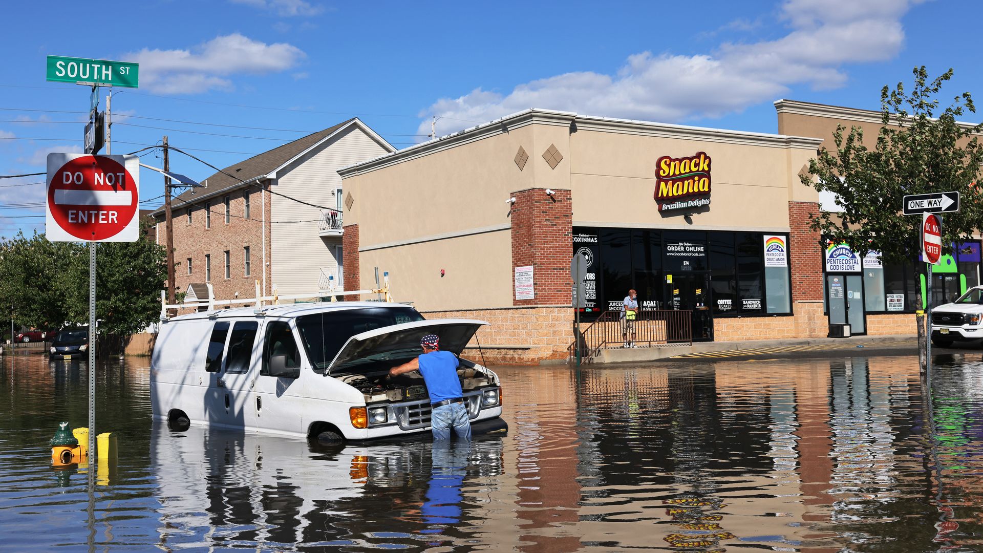man in need deep water works in hood of partially submerged van in flooded intersection.