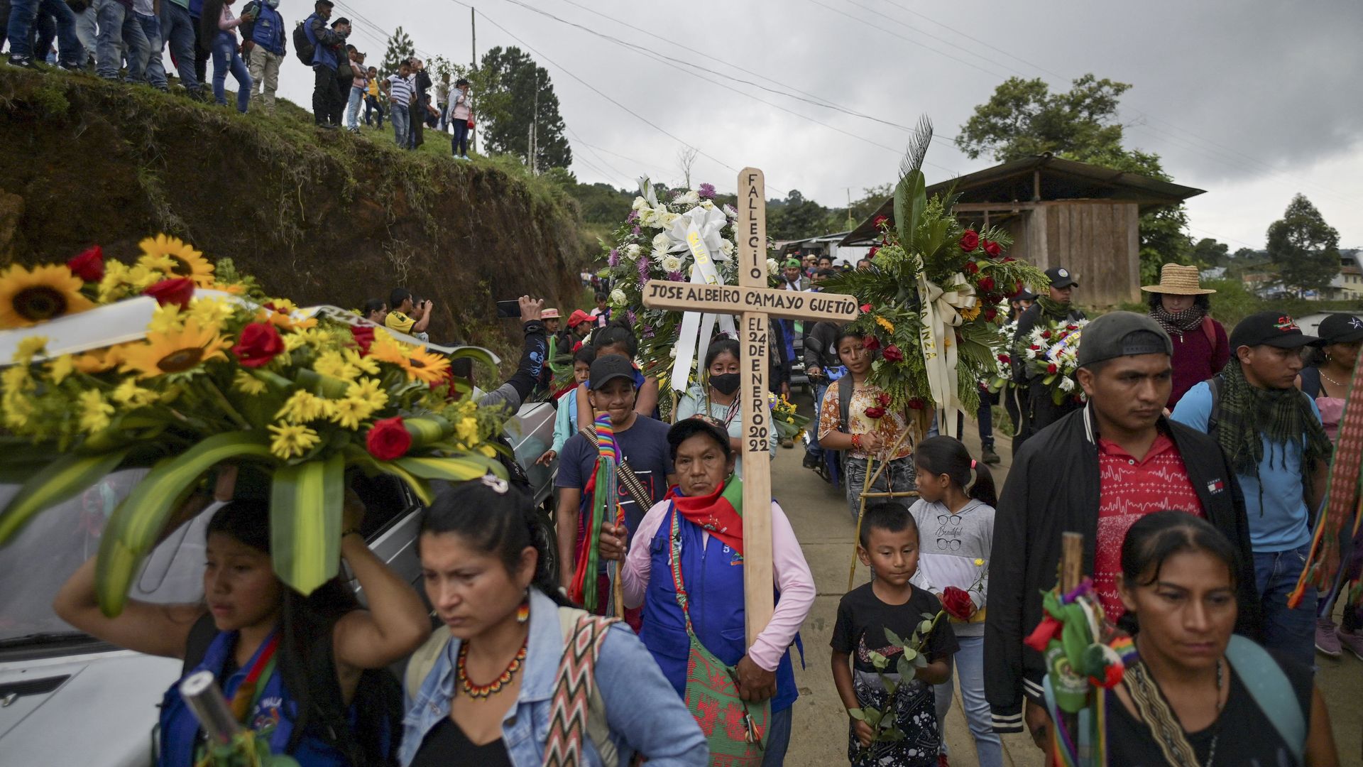 A funeral procession in Colombia in which one woman is carrying a wooden cross, several others are carrying flowers and a child is carrying a red rose